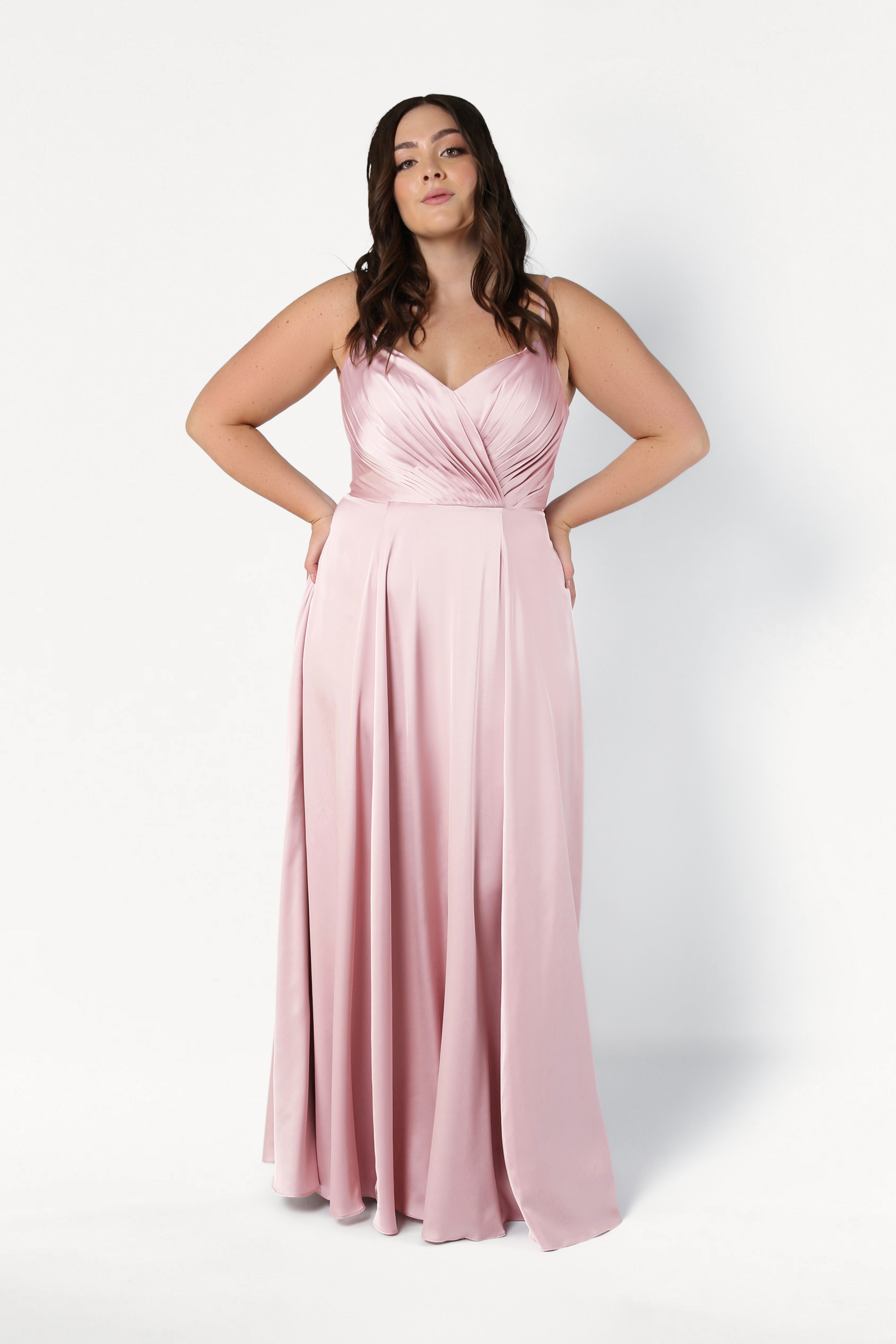 A woman wears the TH&TH Isla Bridesmaid Dress in blush pink satin, featuring a pleated bodice and spaghetti straps, as she stands confidently against a white background with her hands at her hips.