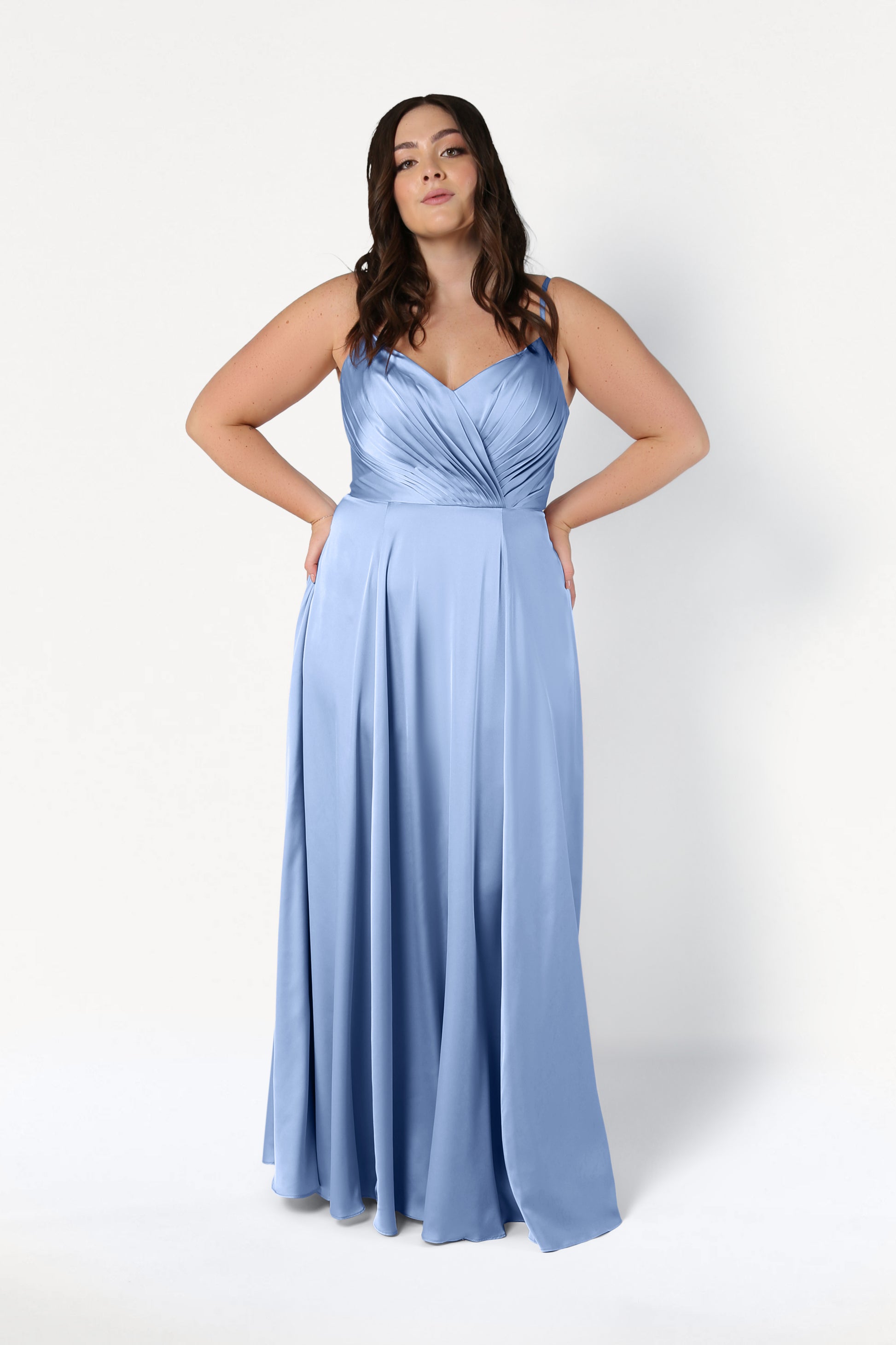 A woman poses in the Isla Bridesmaid Dress in Dusty Blue Satin by TH&TH—a long satin dress with thin straps and a pleated bodice—against a plain white background.