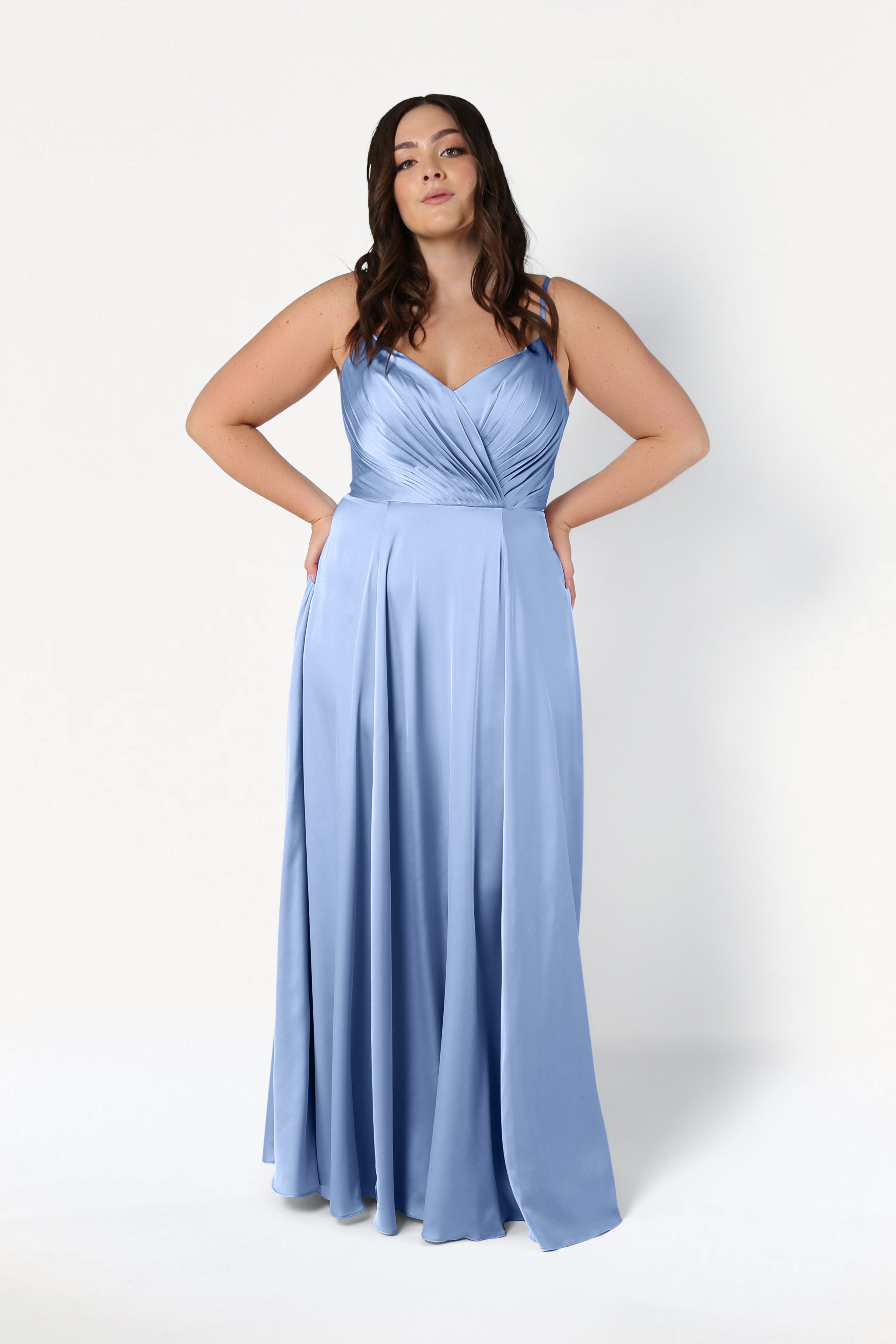 A woman poses in the Isla Bridesmaid Dress in Dusty Blue Satin by TH&TH—a long satin dress with thin straps and a pleated bodice—against a plain white background.