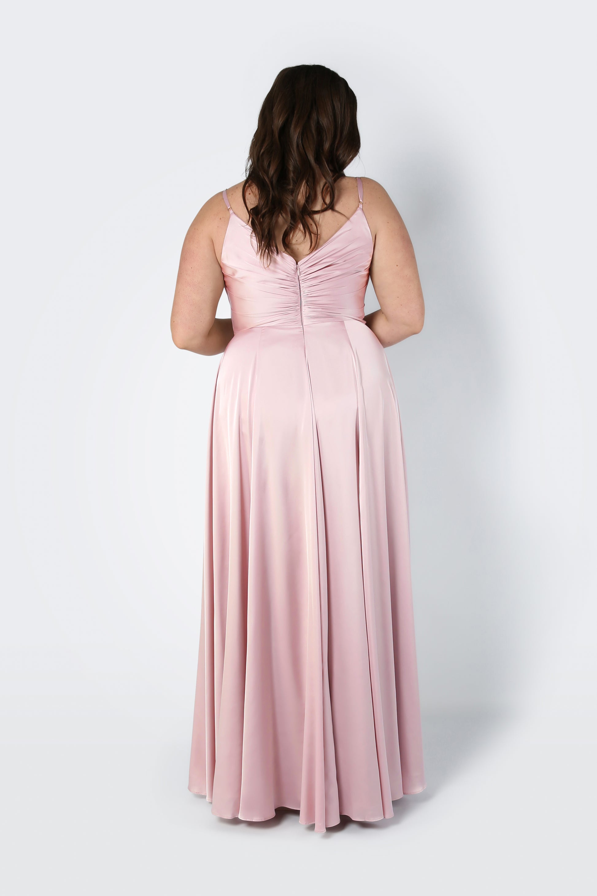 A woman with wavy brown hair faces away from the camera, modeling the TH&TH Isla Bridesmaid Dress in Blush Pink Satin—a sleeveless, floor-length gown with a pleated bodice—against a plain light background.