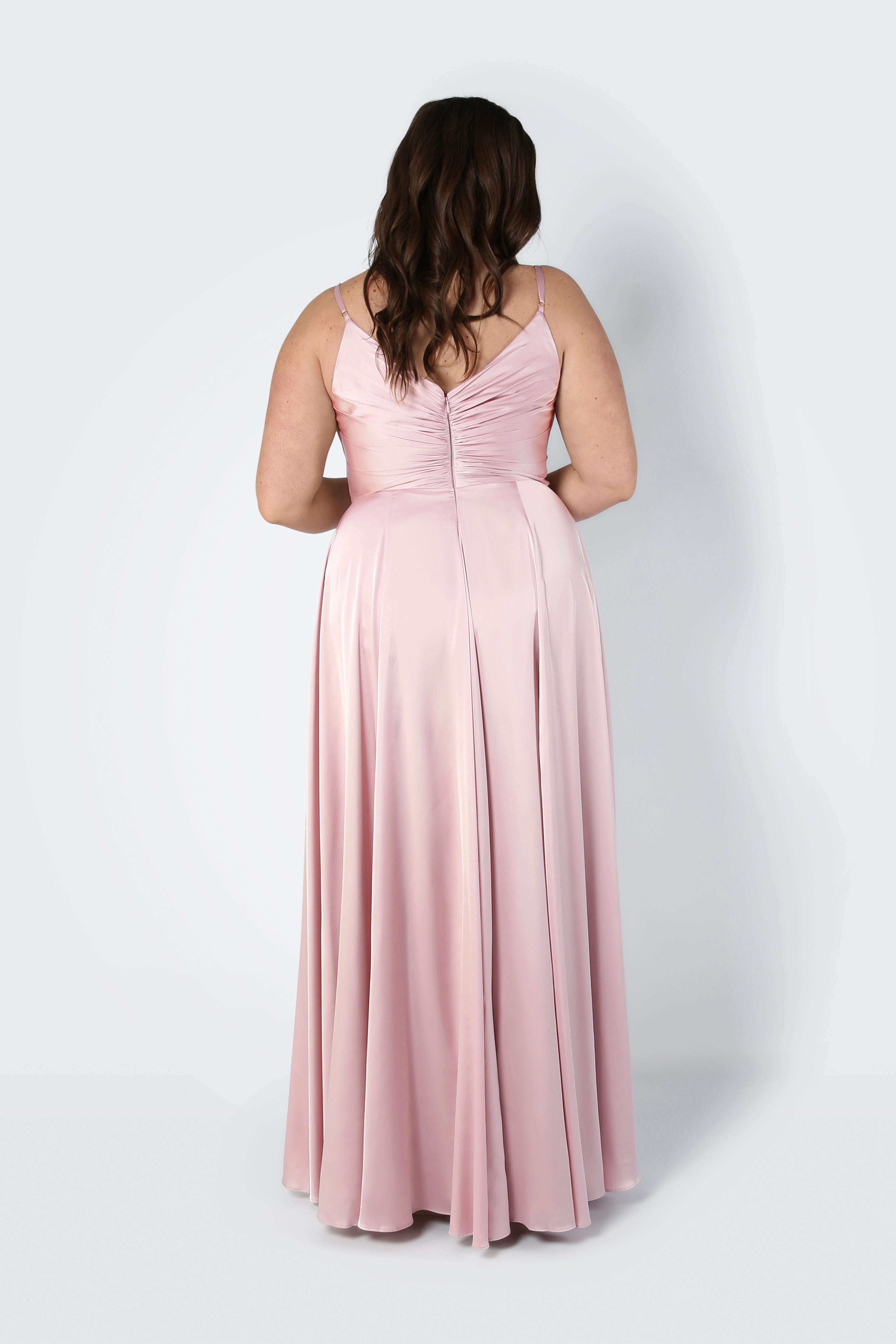 A woman with wavy brown hair faces away from the camera, modeling the TH&TH Isla Bridesmaid Dress in Blush Pink Satin—a sleeveless, floor-length gown with a pleated bodice—against a plain light background.