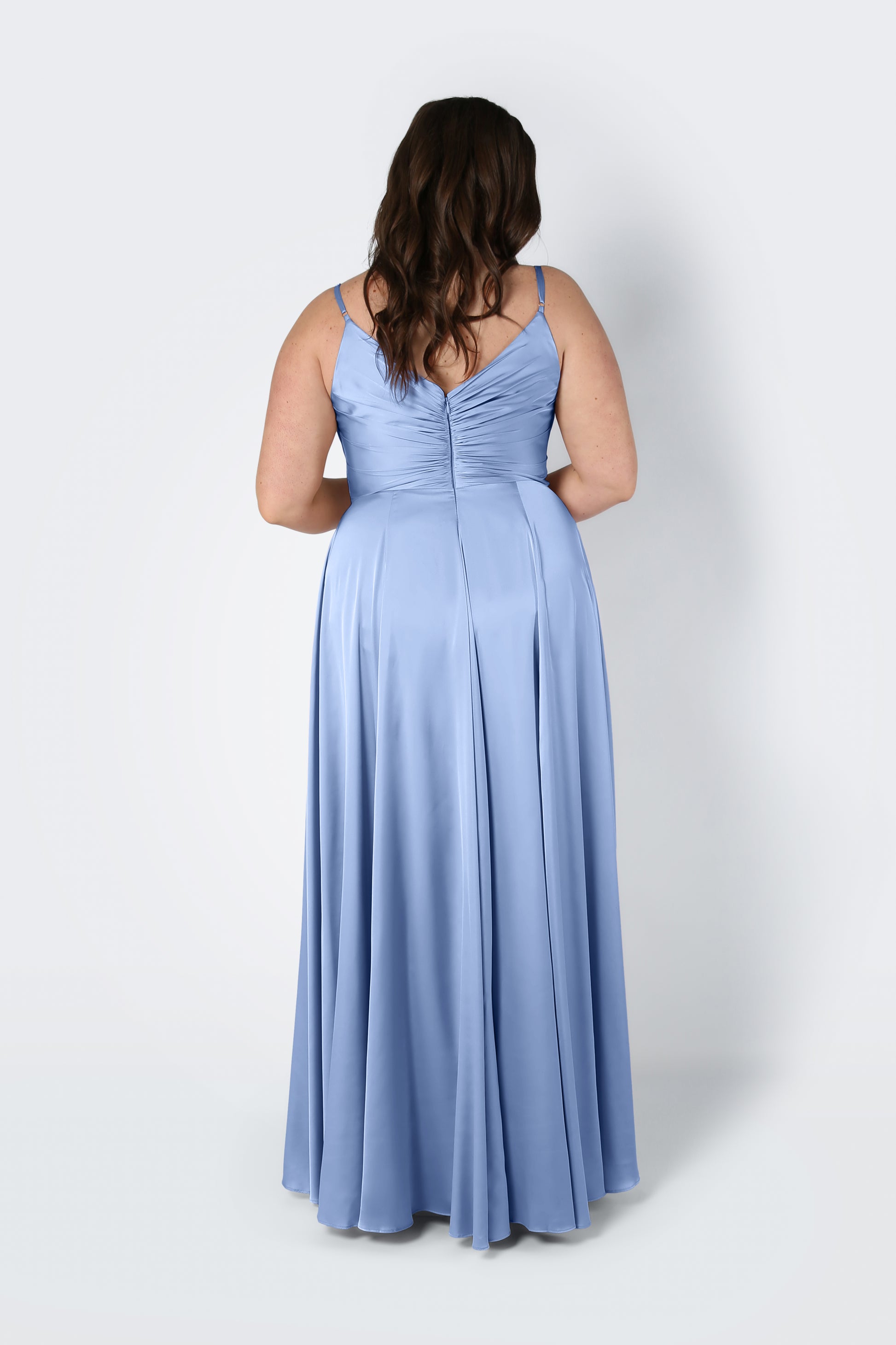 A woman with wavy brown hair models the TH&TH Isla Bridesmaid Dress in Dusty Blue Satin, a long gown with a pleated bodice, spaghetti straps, and ruched back, standing facing away against a plain white background.