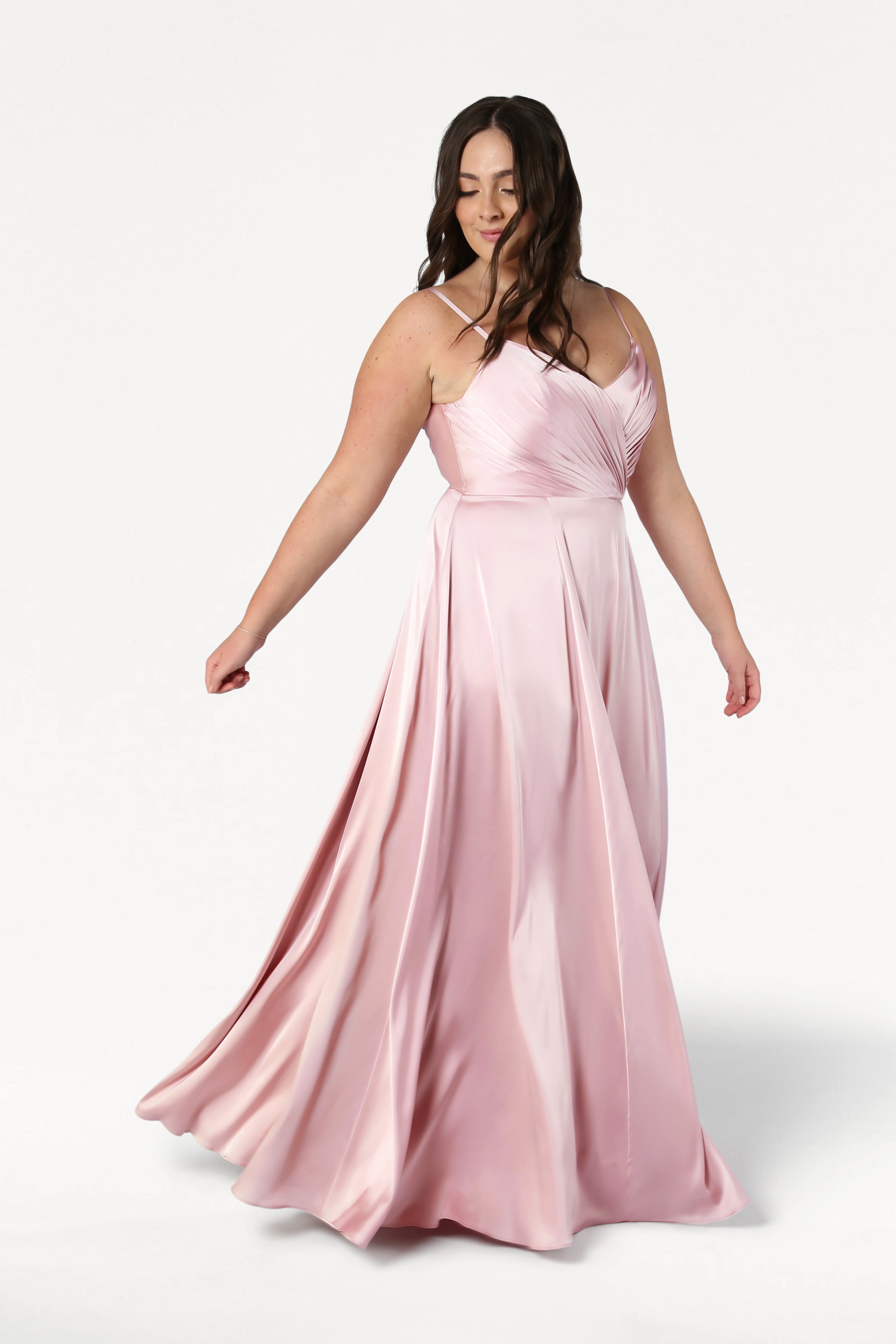 A woman with long brown hair twirls gracefully in the TH&TH Isla Bridesmaid Dress in Blush Pink Satin, featuring a flowing silhouette, pleated bodice, and thin straps, set against a plain white background.