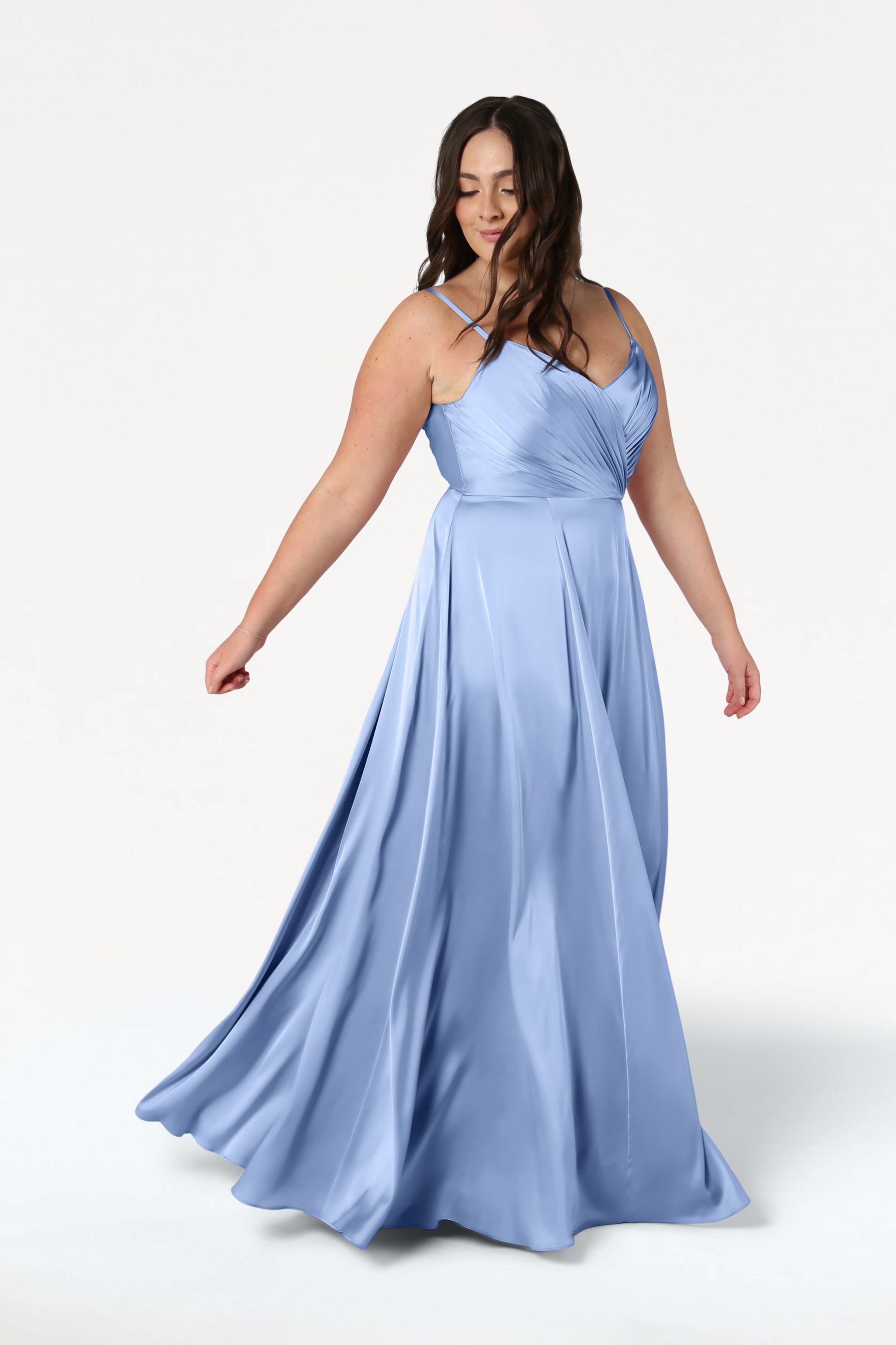 A woman twirls gracefully in the Isla Bridesmaid Dress in Dusty Blue Satin by TH&TH, featuring a pleated bodice and thin straps, smiling gently against a plain white background.