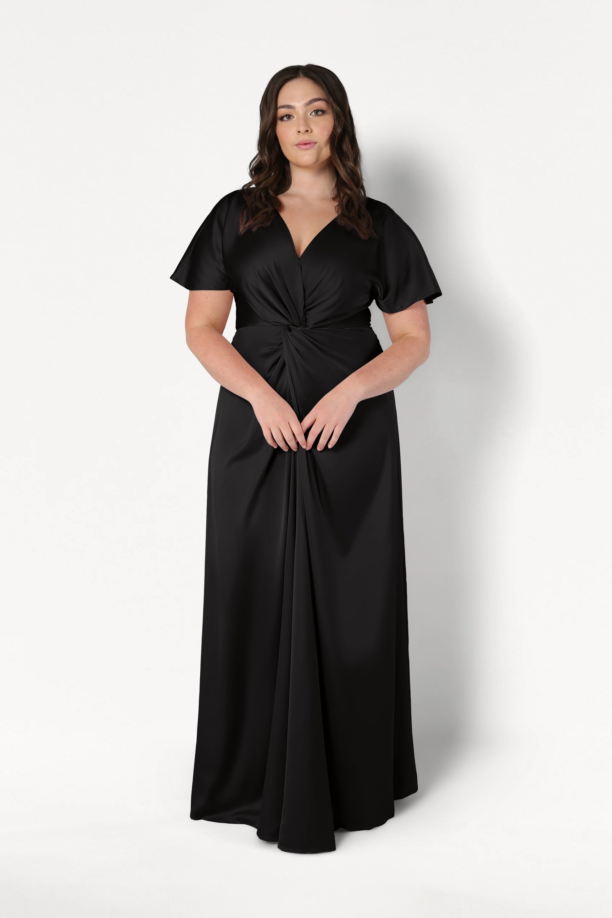 A woman wears the Camilla Bridesmaid Dress in Black Satin by TH&TH—a floor-length, short-sleeved satin gown with a twist-knot bodice—standing against a plain white background.