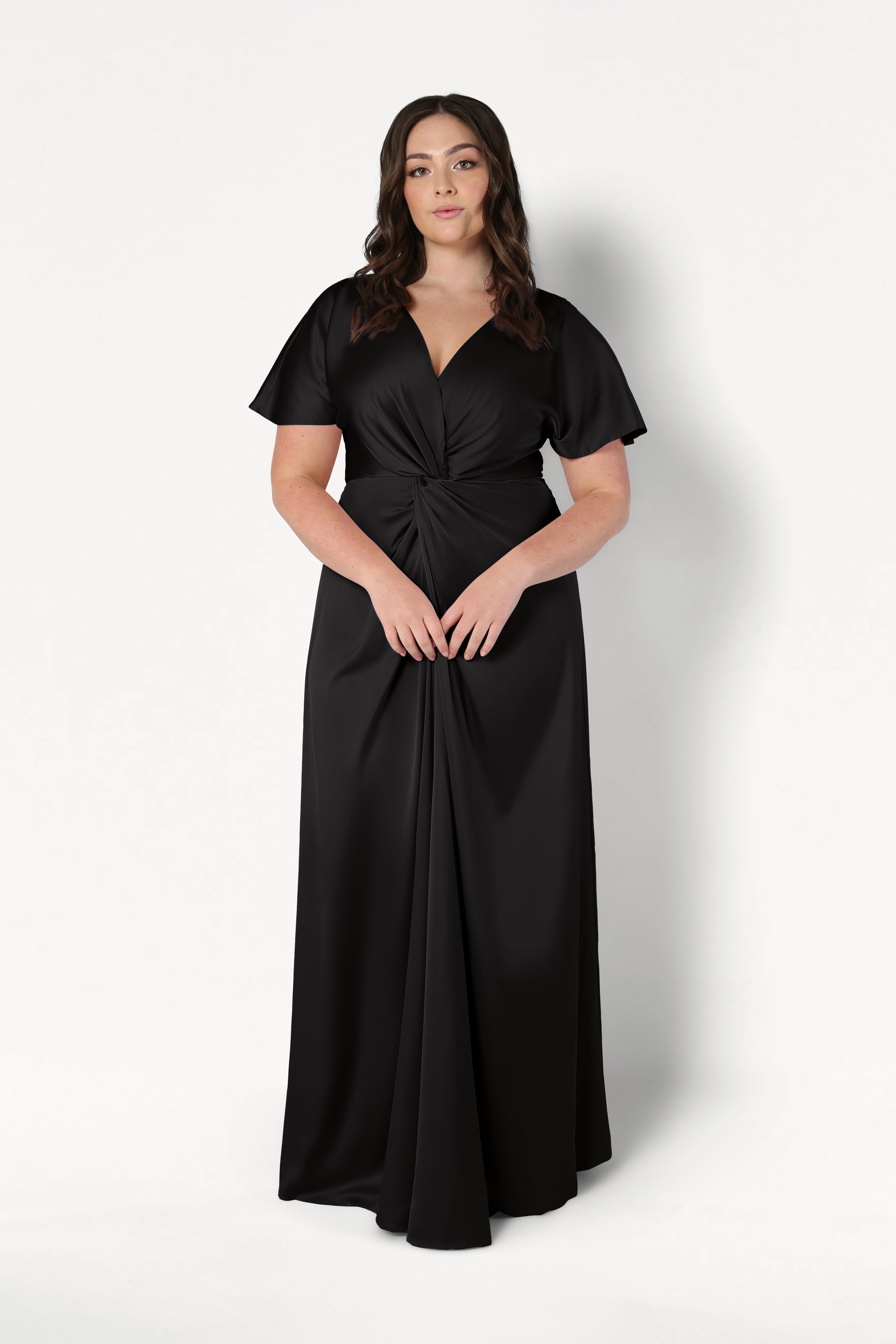 A woman wears the Camilla Bridesmaid Dress in Black Satin by TH&TH—a floor-length, short-sleeved satin gown with a twist-knot bodice—standing against a plain white background.