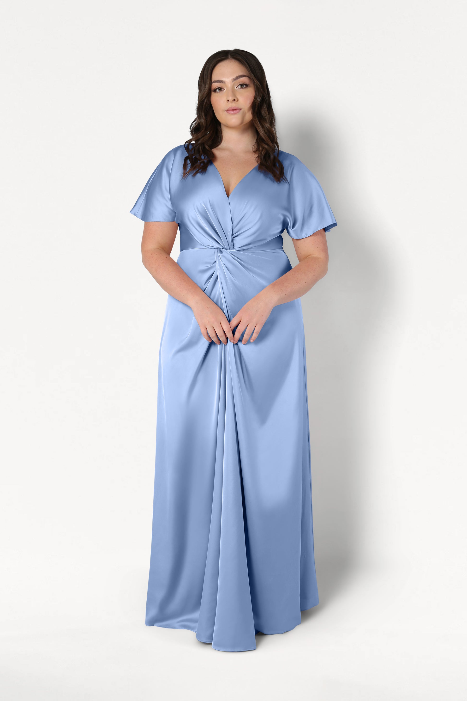 A woman with long, wavy brown hair and a neutral expression stands against a white background, wearing the TH&TH Camilla Bridesmaid Dress in Dusty Blue Satin—a floor-length, short-sleeved gown featuring a knotted waist detail.
