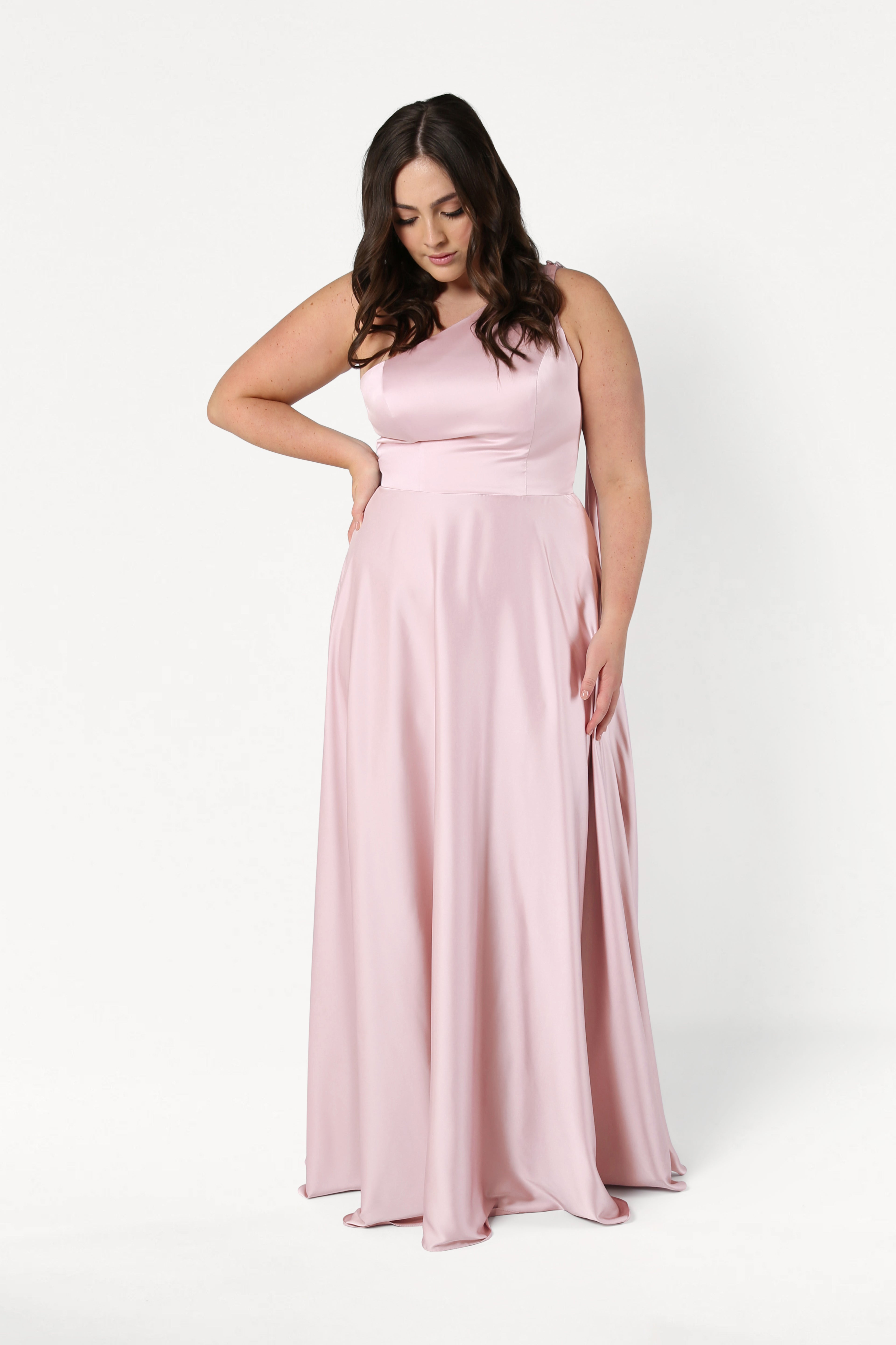 A woman wears the Lyra Bridesmaid Dress in Blush Pink Satin by TH&TH, standing against a white background with one hand on her hip and the other gently touching the dress.