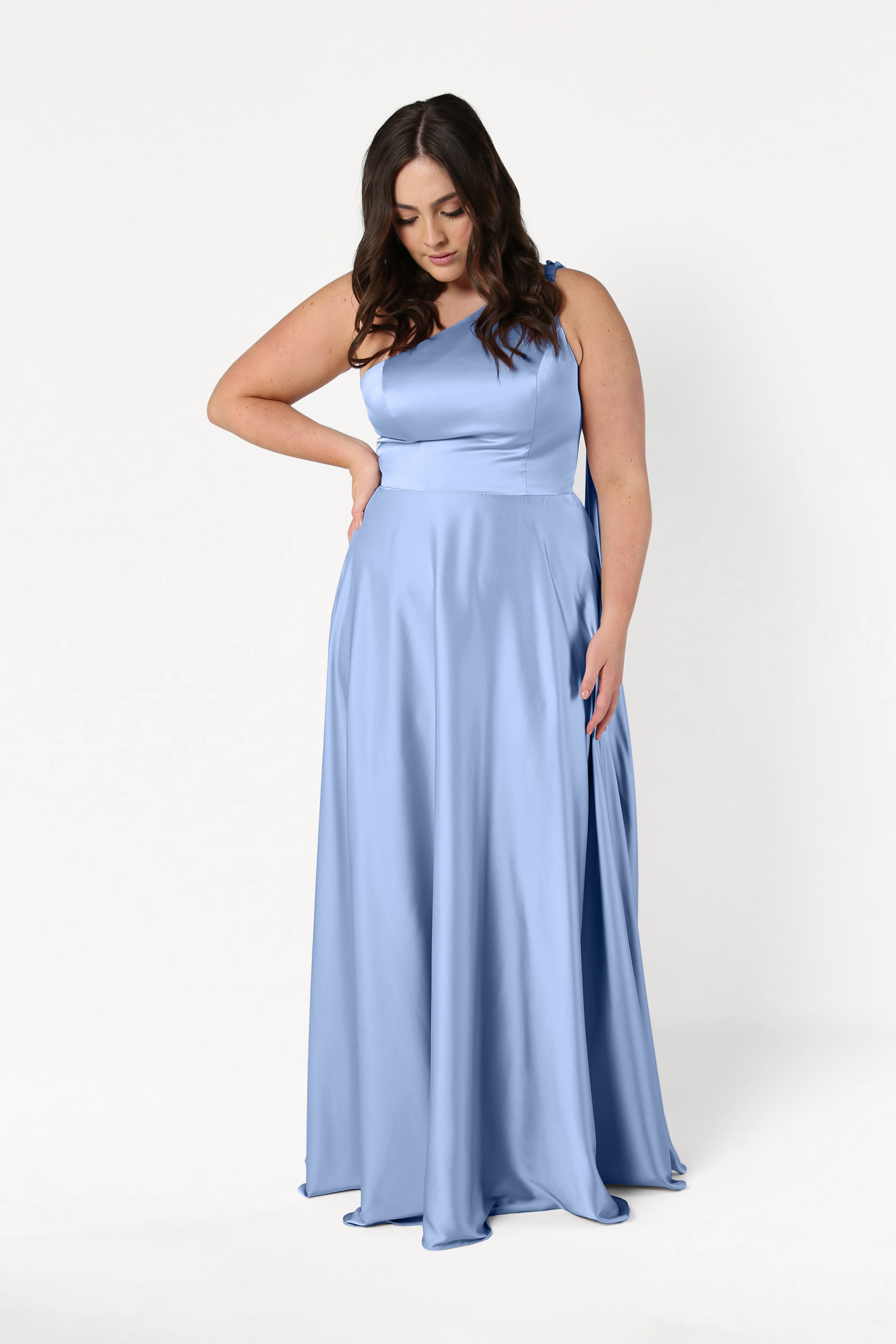 A woman in the Lyra Bridesmaid Dress by TH&TH, crafted in dusty blue satin, stands against a plain white background, looking down with one arm on her waist and the other gently holding her dress.