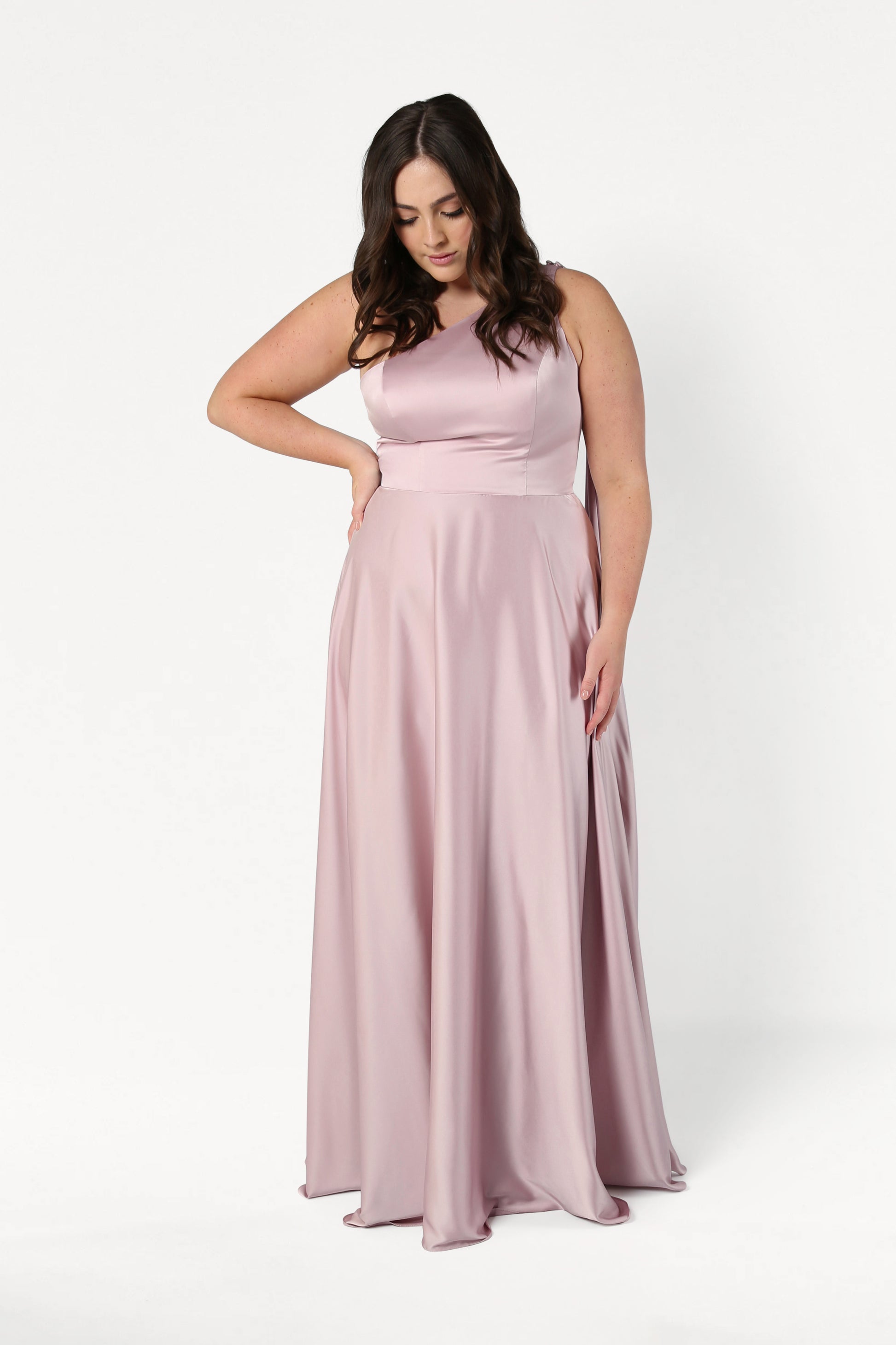 A woman with long dark hair wears the Lyra Bridesmaid Dress in Smoked Blush Satin by TH&TH. This elegant, sleeveless, floor-length gown is size-inclusive. She stands against a white background, looking down with one hand on her hip.