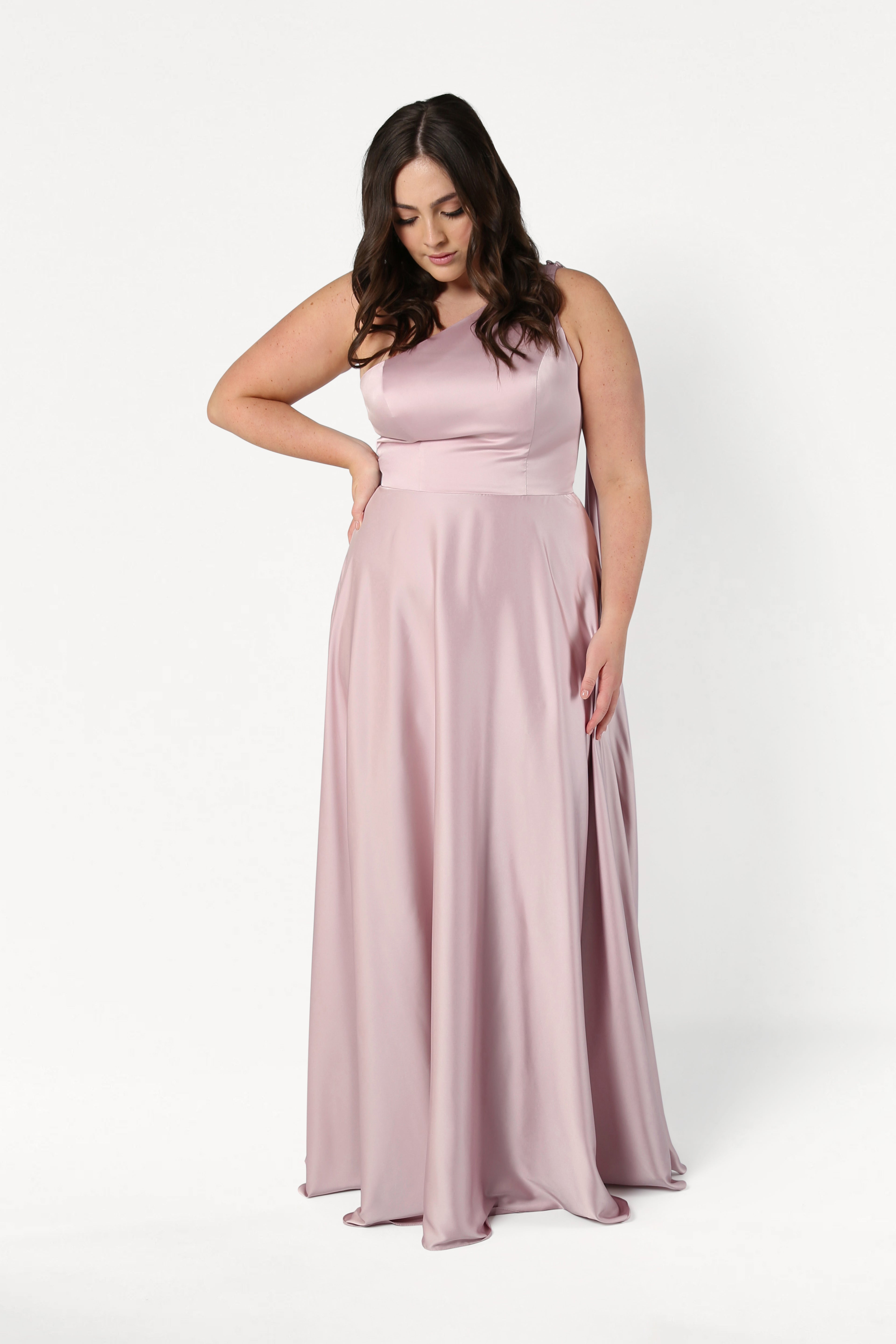 A woman with long dark hair wears the Lyra Bridesmaid Dress in Smoked Blush Satin by TH&TH. This elegant, sleeveless, floor-length gown is size-inclusive. She stands against a white background, looking down with one hand on her hip.