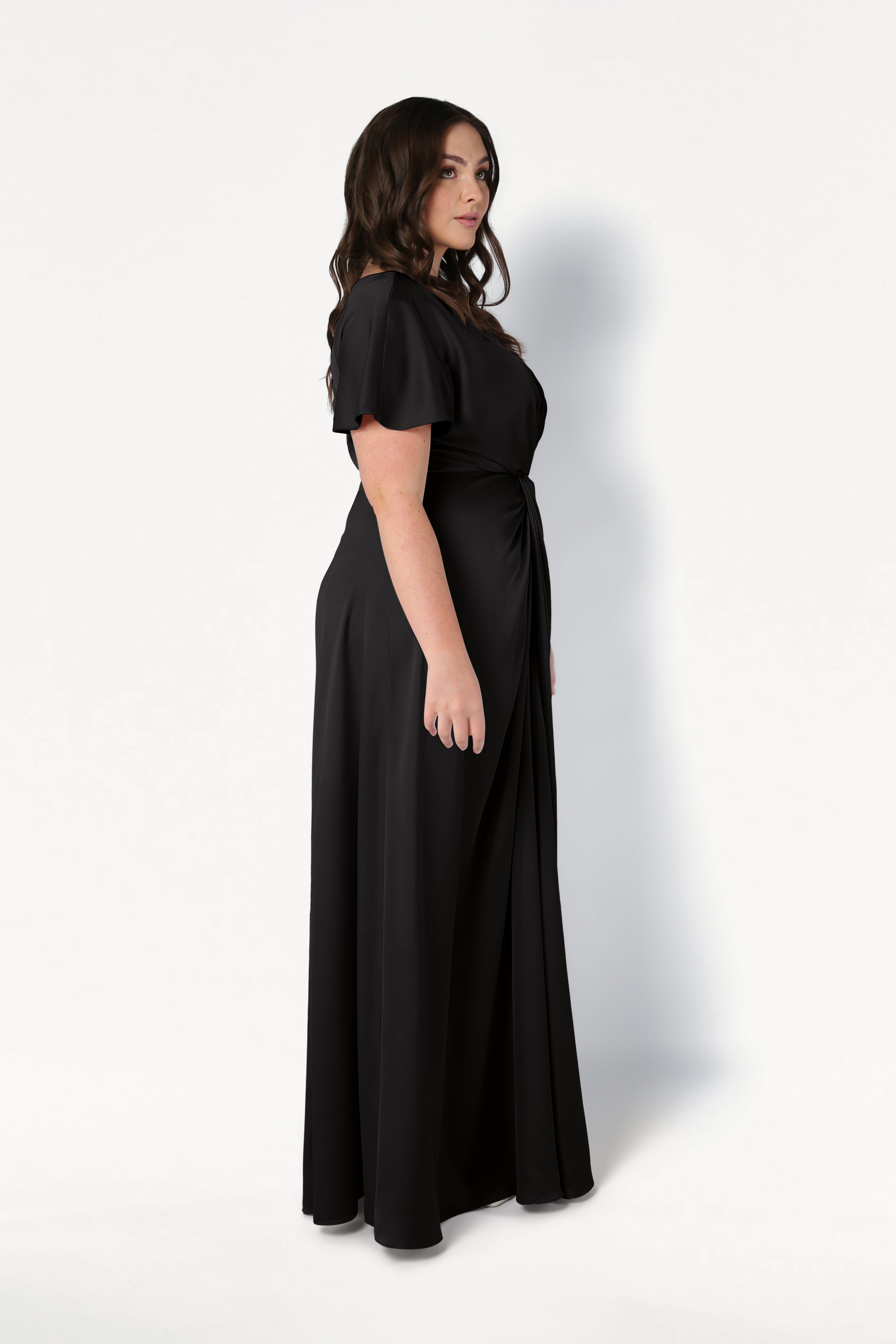 A woman with long wavy hair stands sideways in the TH&TH Camilla Bridesmaid Dress in black satin, a size-inclusive, floor-length gown with short sleeves and a gathered waist, set against a plain white background.