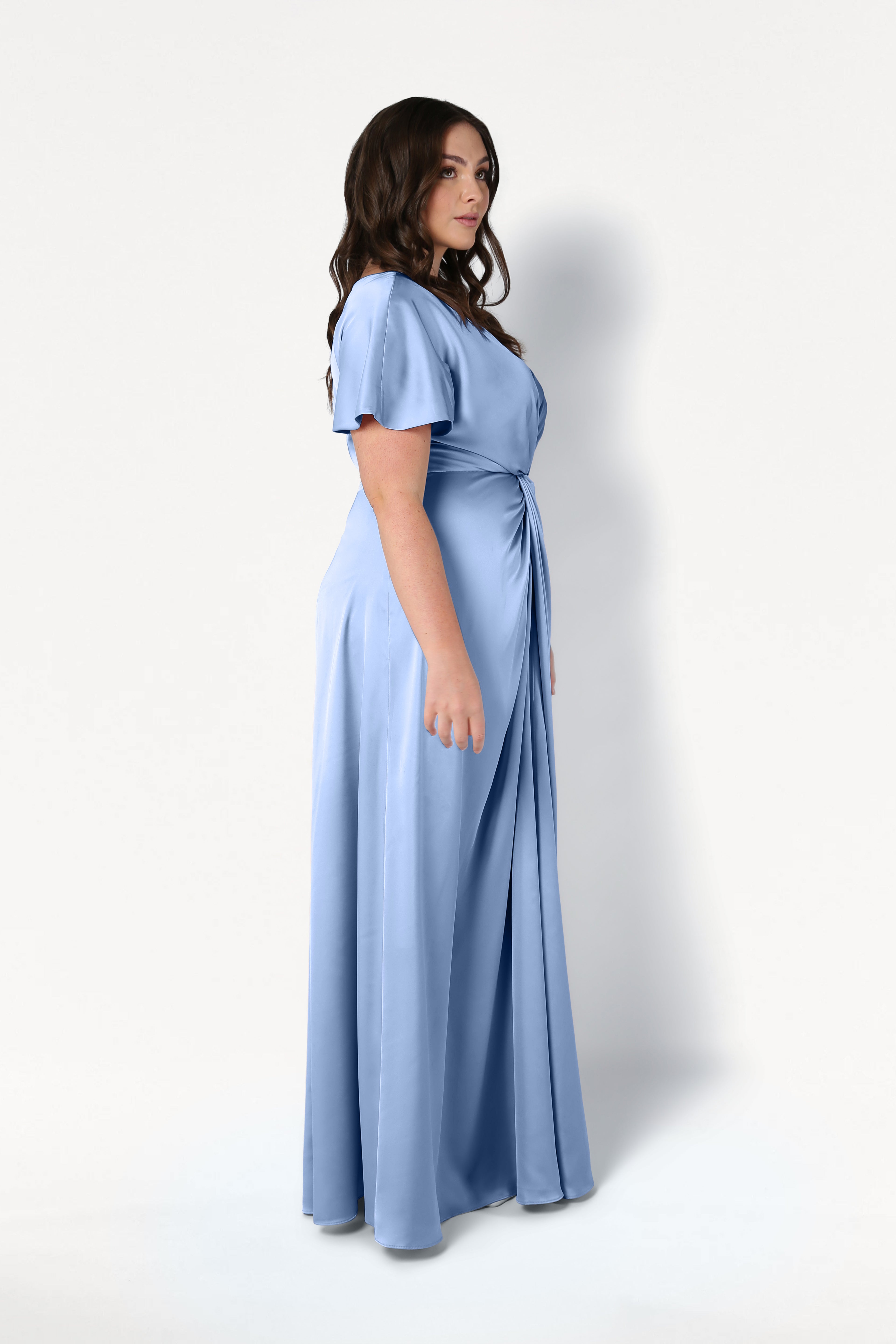 A woman with long dark hair stands sideways, modeling the TH&TH Camilla Bridesmaid Dress in Dusty Blue Satin—a size-inclusive, floor-length gown with gentle draping and a gathered waist—against a plain white background.