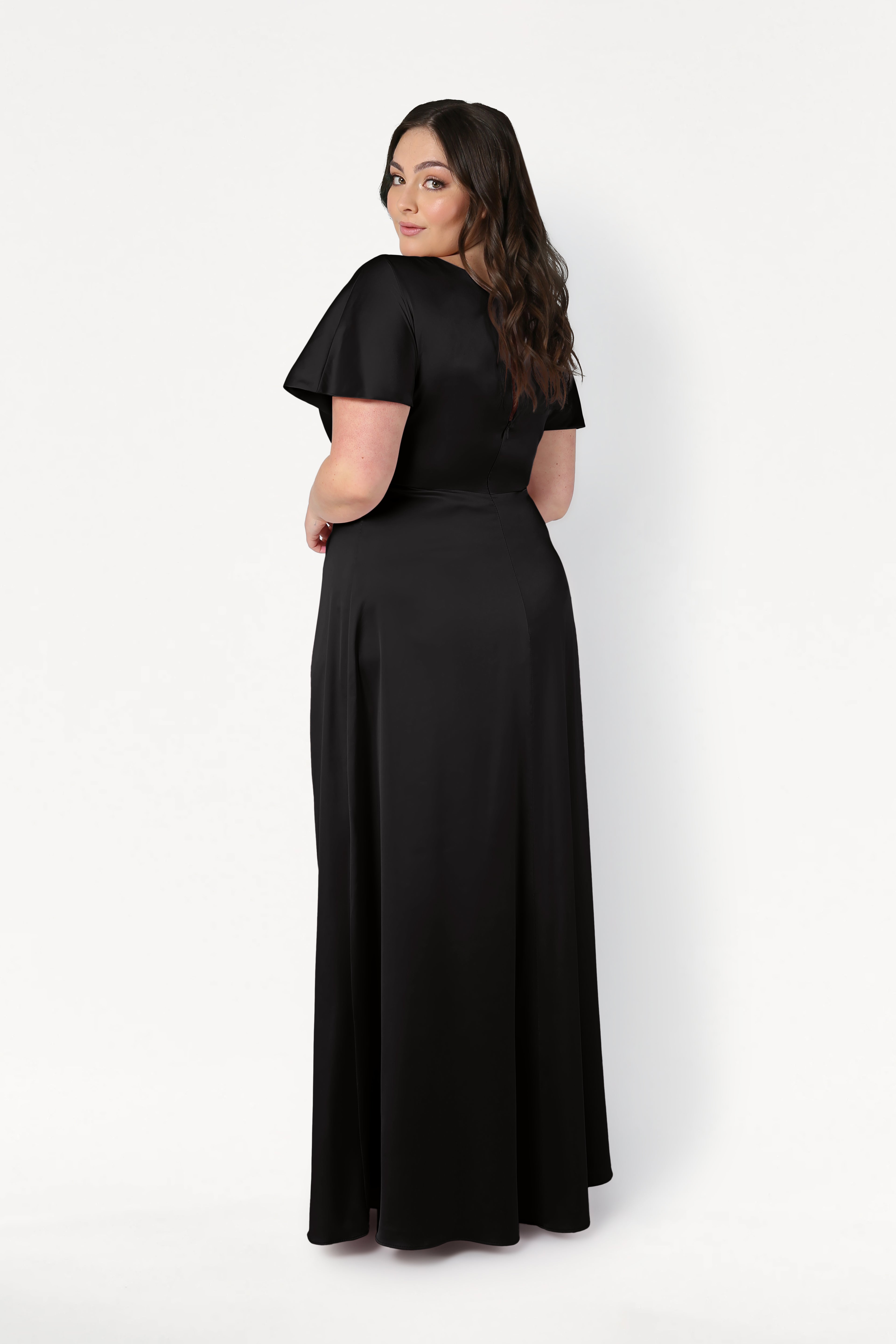 A woman with long dark hair, facing away and looking over her shoulder, models the size-inclusive TH&TH Camilla Bridesmaid Dress in Black Satin—a floor-length gown with a twist-knot bodice—against a plain white background.