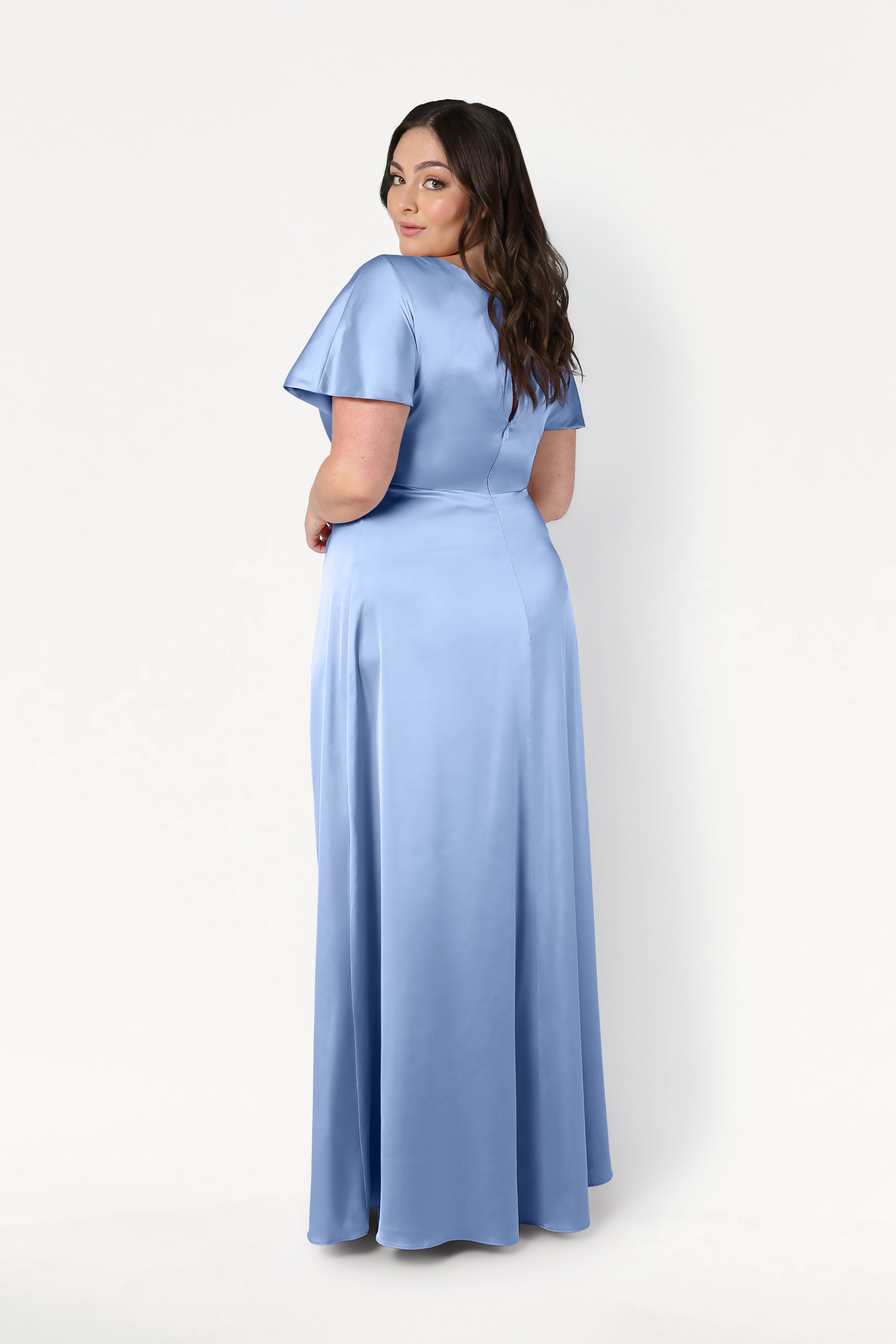 A woman with long brown hair models the Camilla Bridesmaid Dress in Dusty Blue Satin by TH&TH, featuring short sleeves and a floor-length design, as she looks over her shoulder against a plain white background.