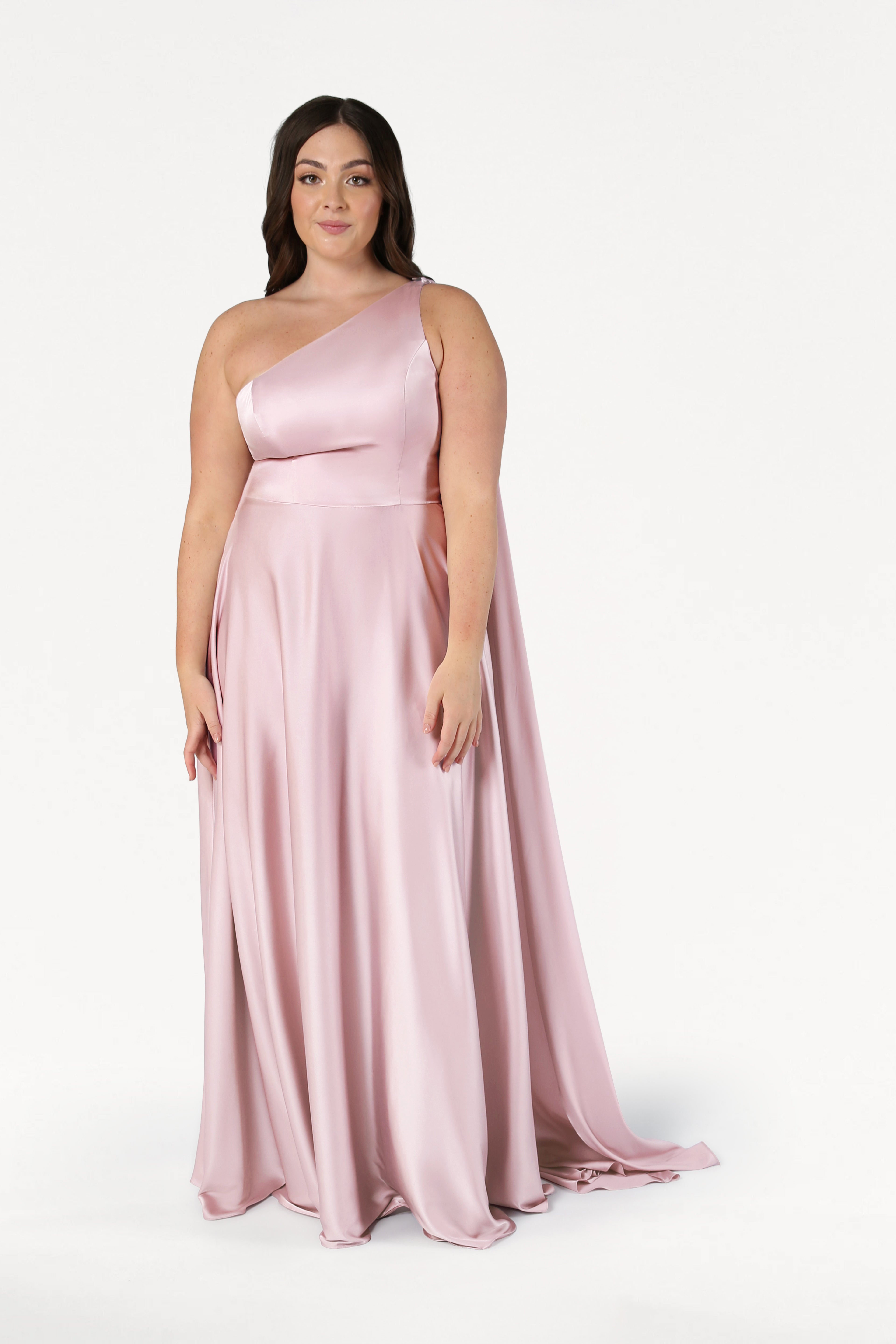 A woman models the TH&TH Lyra Bridesmaid Dress in blush pink satin, featuring a long, flowing silhouette and one-shoulder design, standing against a plain white background and looking at the camera.