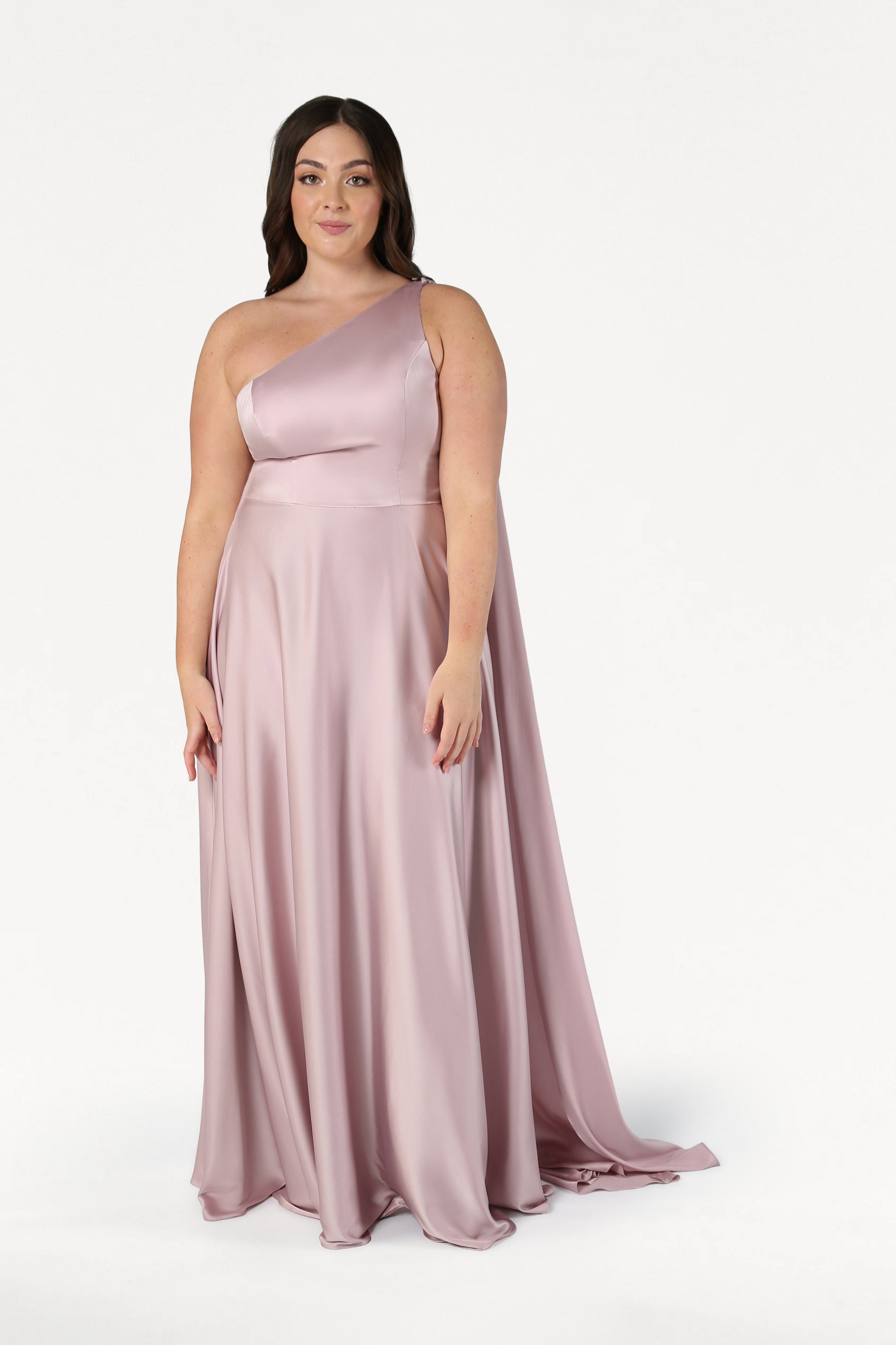 A woman in the Lyra Bridesmaid Dress in Smoked Blush Satin by TH&TH poses against a white background, softly smiling. This elegant, size-inclusive one-shoulder gown is perfect for every celebration.