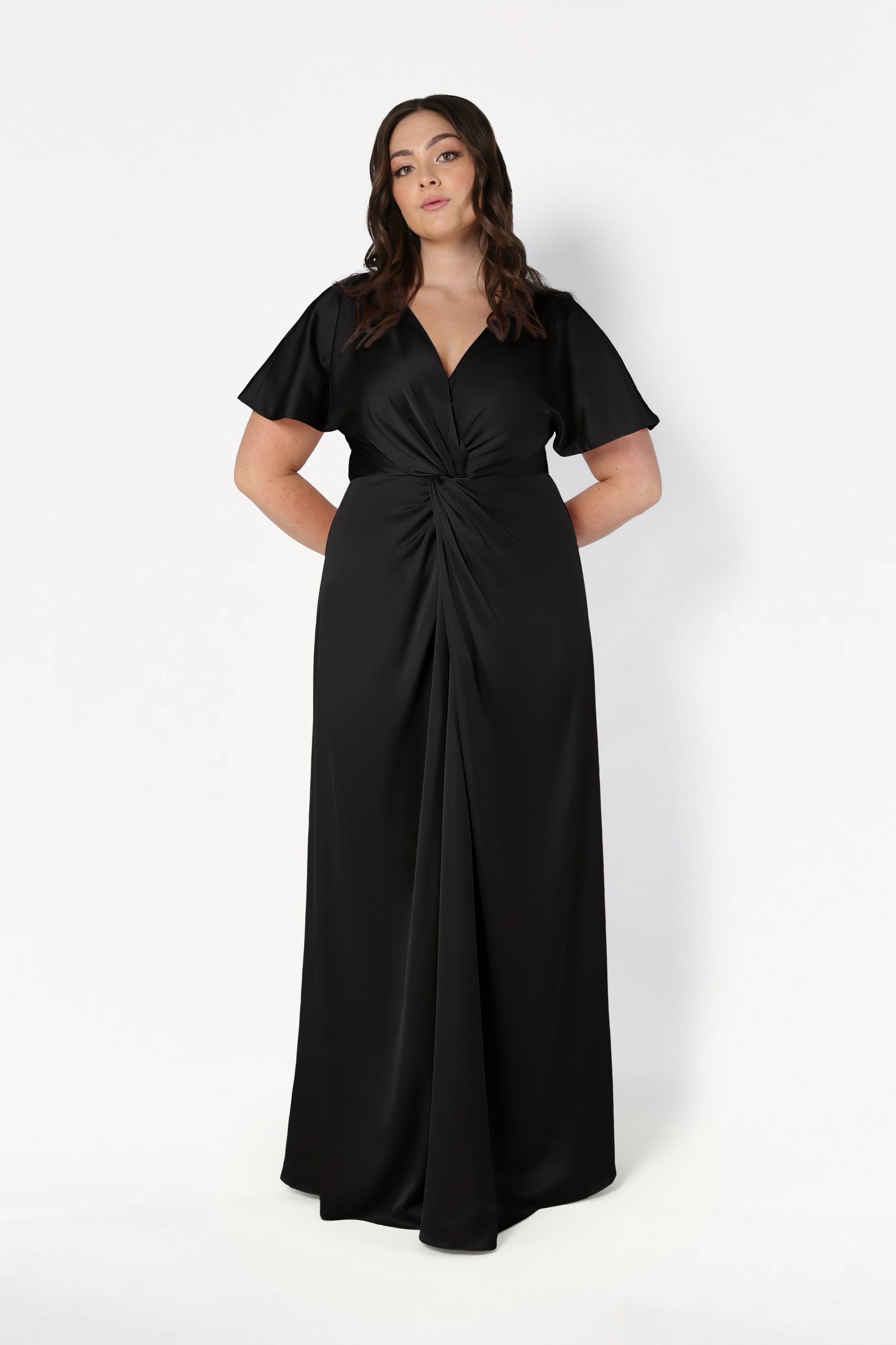 A woman models the Camilla Bridesmaid Dress in Black Satin by TH&TH, featuring a floor-length silhouette, short sleeves, V-neckline, and twist-knot bodice, standing confidently against a white background.