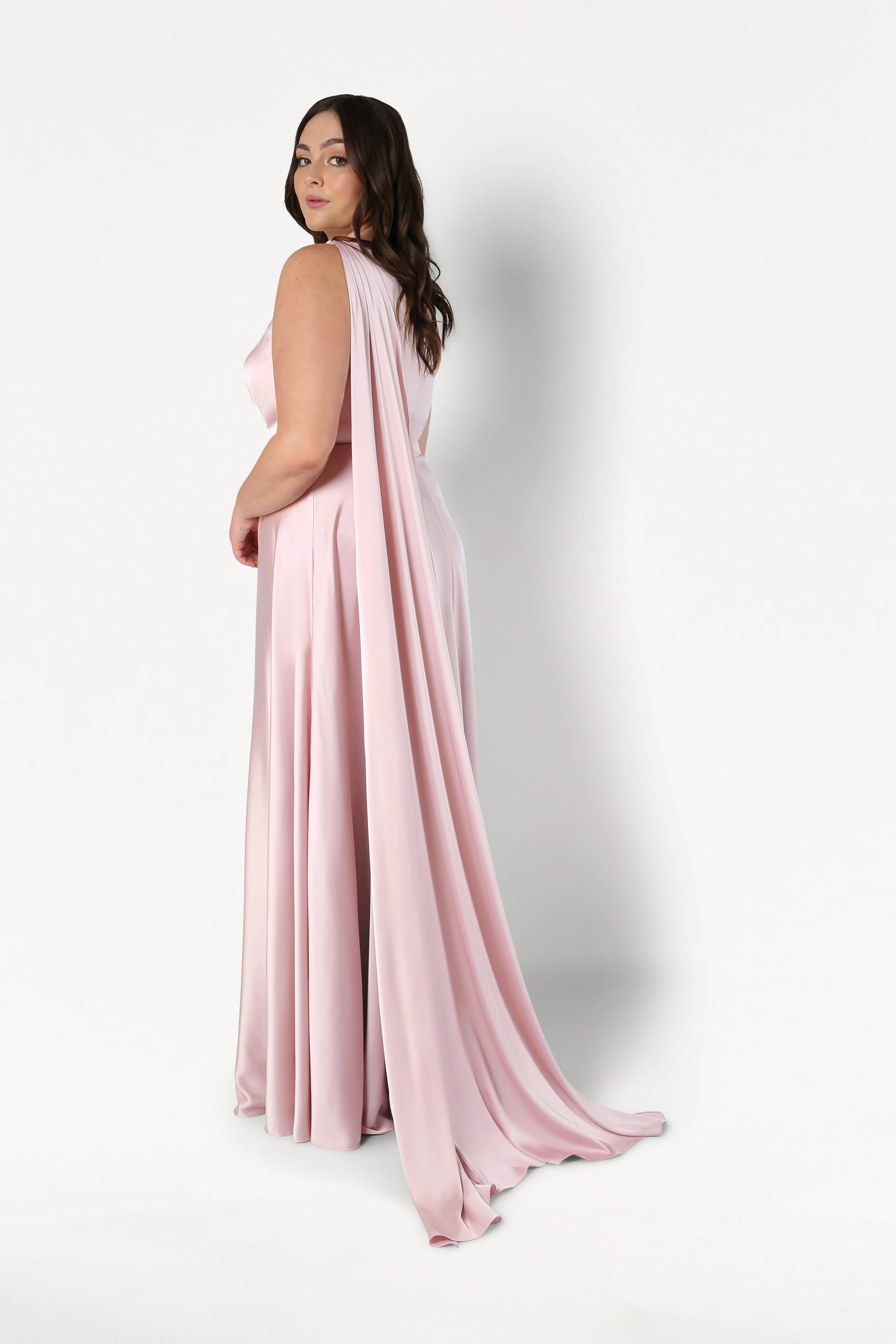 A woman with wavy brown hair models the TH&TH Lyra Bridesmaid Dress in blush pink satin, featuring a size-inclusive fit and long, flowing cape, as she looks over her shoulder against a plain white background.
