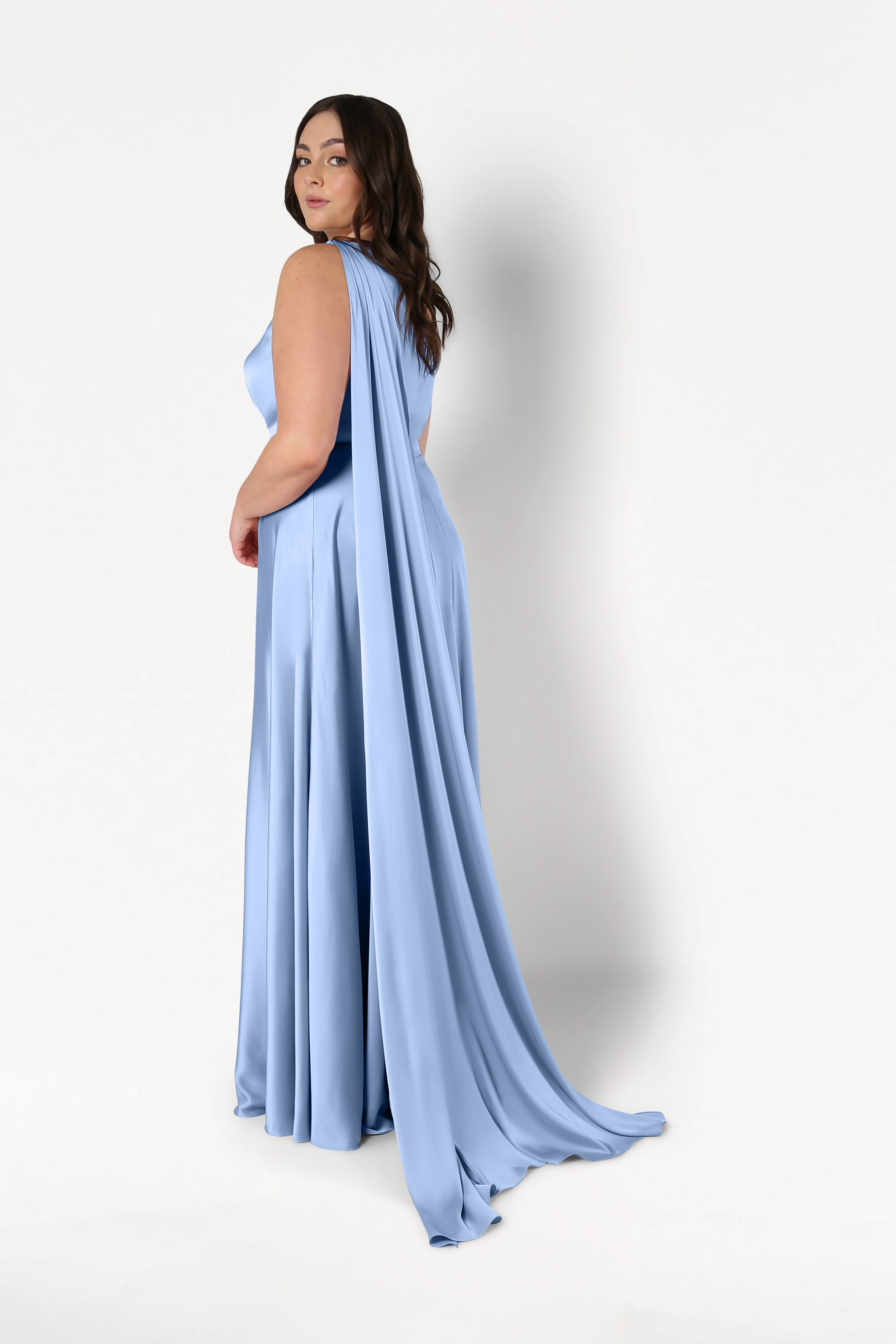 A woman models the Lyra Bridesmaid Dress in Dusty Blue Satin by TH&TH, featuring a flowing, size-inclusive design with a long draped cape, as she poses against a plain white background looking over her shoulder.