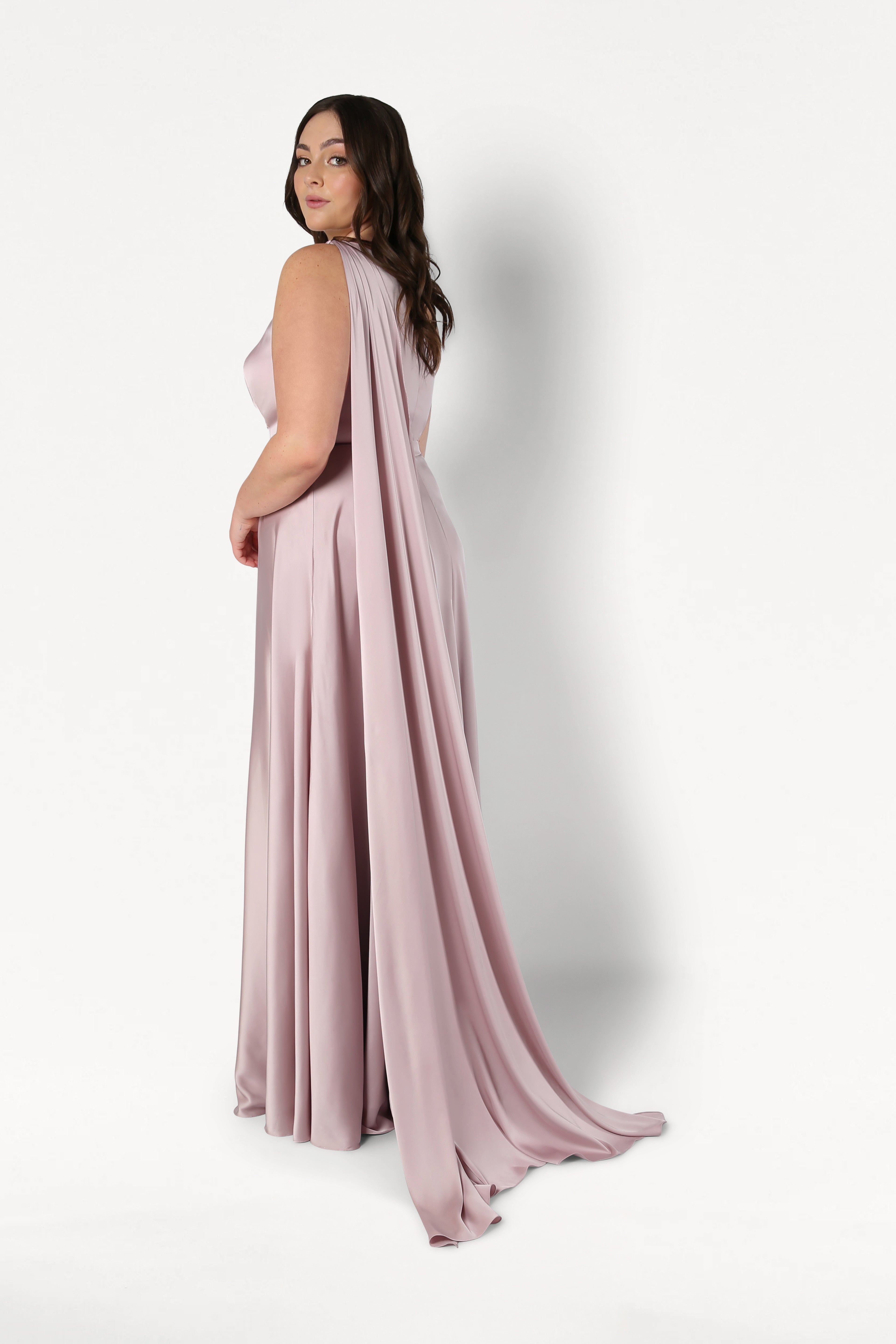 A woman in the TH&TH Lyra Bridesmaid Dress in Smoked Blush Satin stands against a white background, glancing over her shoulder with wavy hair. The size-inclusive, draped-back style flatters every silhouette.