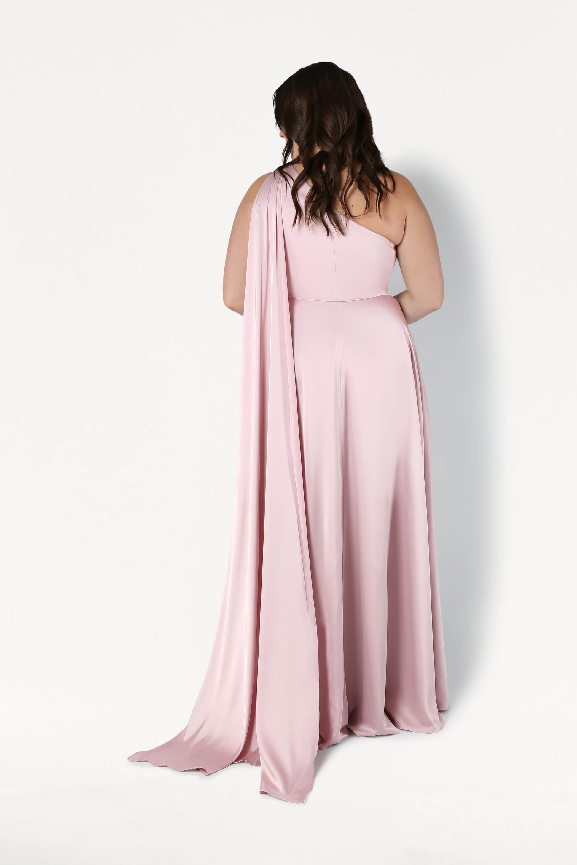 A woman with wavy brown hair faces away, wearing the TH&TH Lyra Bridesmaid Dress in blush pink satin—a size-inclusive, one-shoulder gown with flowing draped fabric—set against a plain white background.
