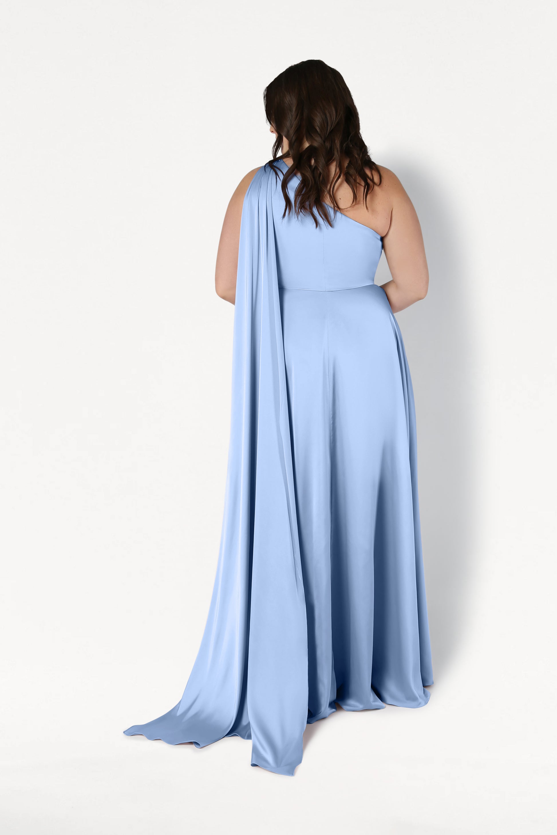 A woman with wavy brown hair stands with her back to the camera, wearing the TH&TH Lyra Bridesmaid Dress in Dusty Blue Satin—a floor-length, one-shoulder gown with a draped sash—shown against a plain white background.