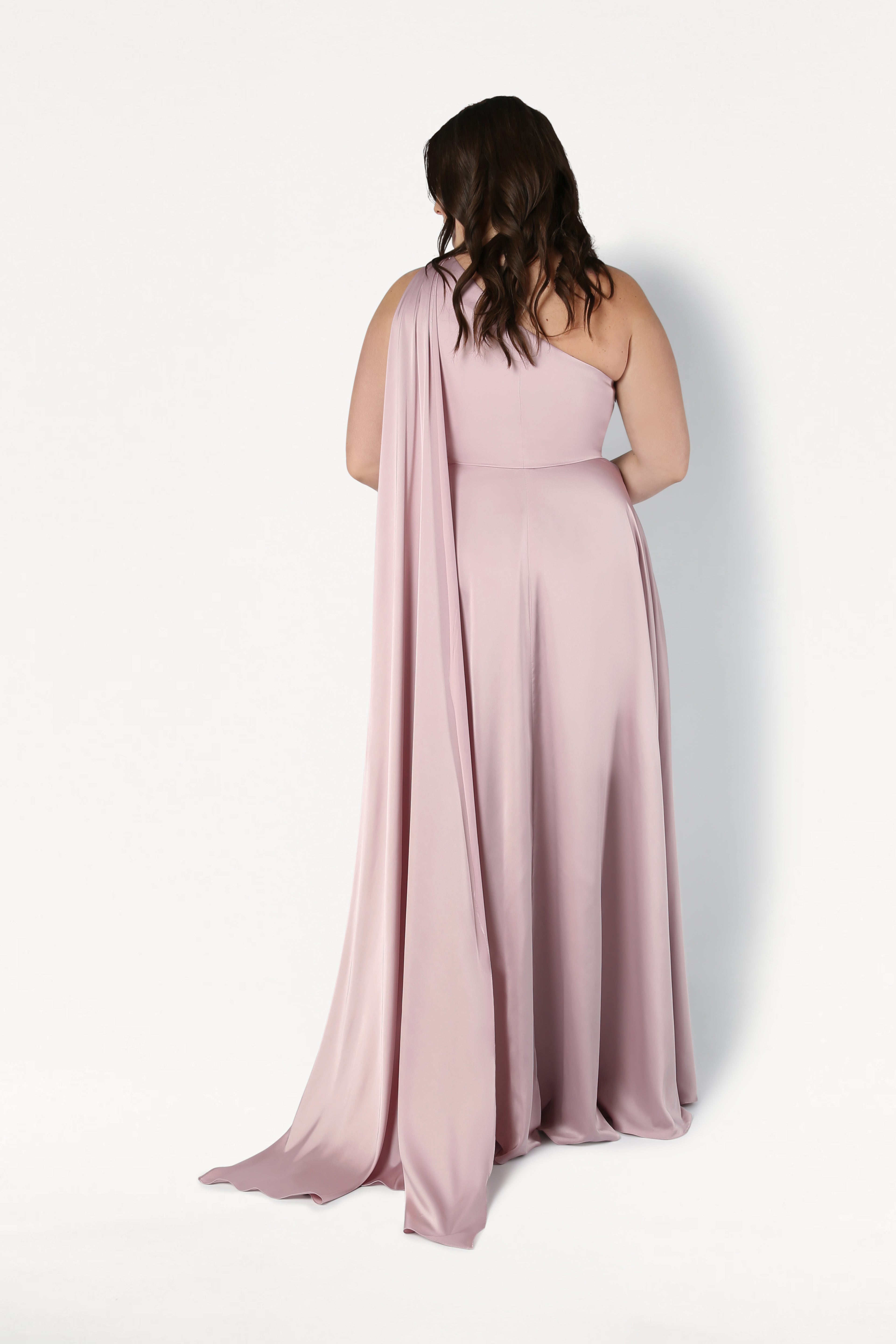 A woman with wavy brown hair stands with her back to the camera, wearing the size-inclusive Lyra Bridesmaid Dress in Smoked Blush Satin by TH&TH. The one-shoulder satin gown flows elegantly, featuring draped fabric against a plain white background.