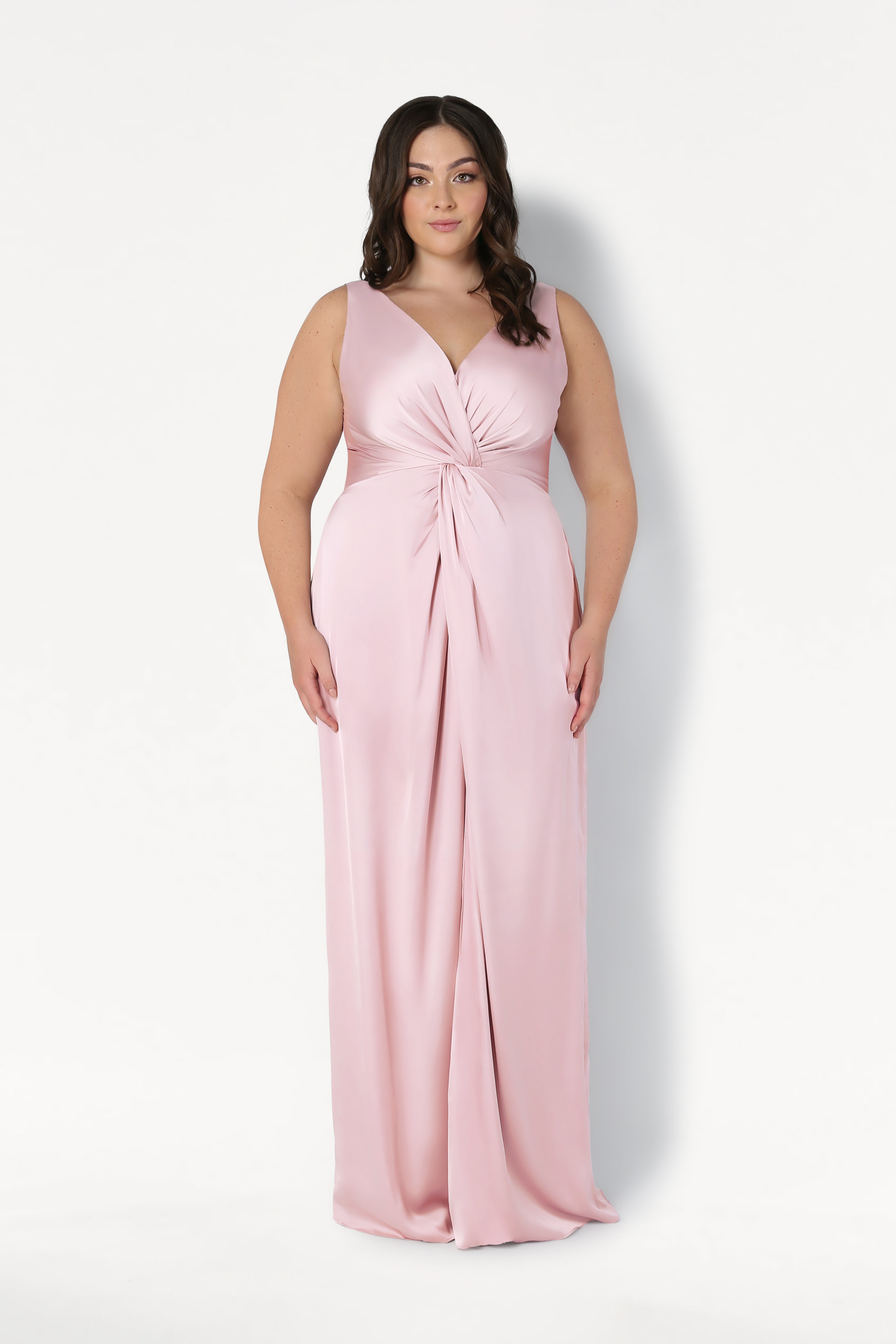 A woman with long, dark wavy hair models the TH&TH Carmen Bridesmaid Dress in Blush Pink Satin, a sleeveless gown with a twist-knot waist detail, standing before a plain white background.