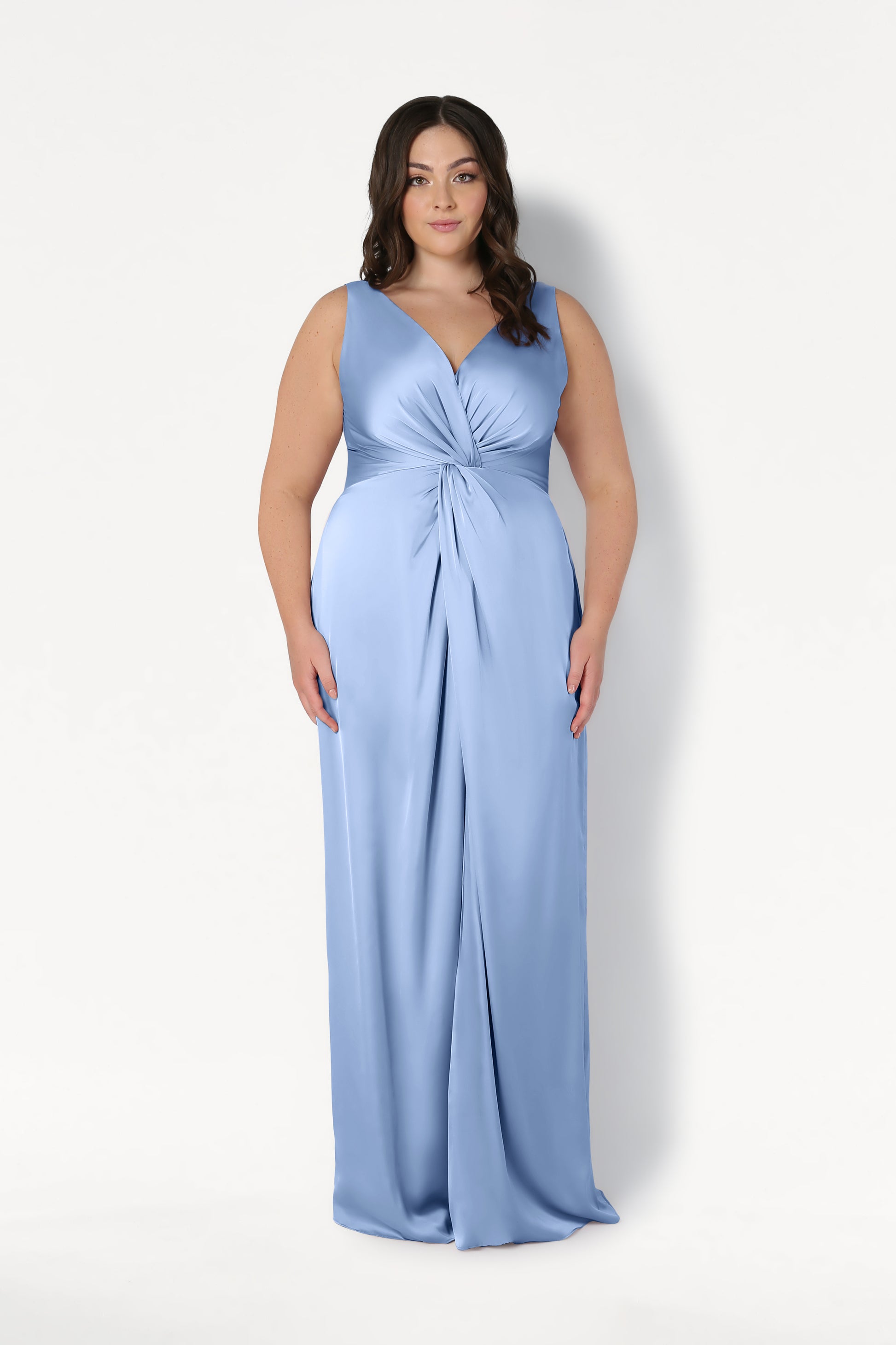 A woman stands against a white background wearing the Carmen Bridesmaid Dress in Dusty Blue Satin by TH&TH, featuring a twist-knot waist and V-neckline—perfect for modern bridal parties.