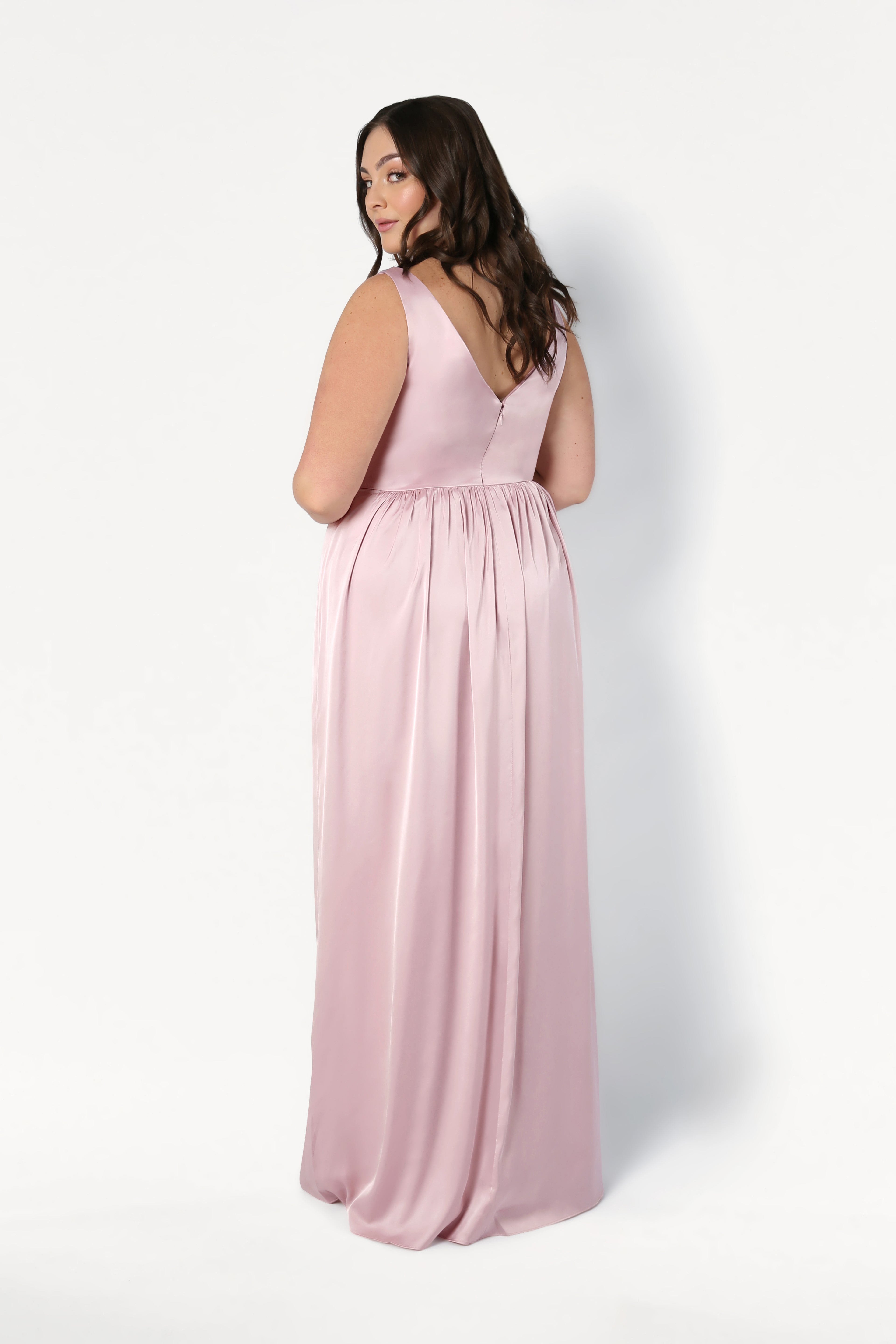 A woman with long brown hair stands against a white background, wearing the TH&TH Carmen Bridesmaid Dress in Blush Pink Satin, a sleeveless floor-length gown featuring a deep V-back. She is turned slightly to the side, looking over her shoulder.