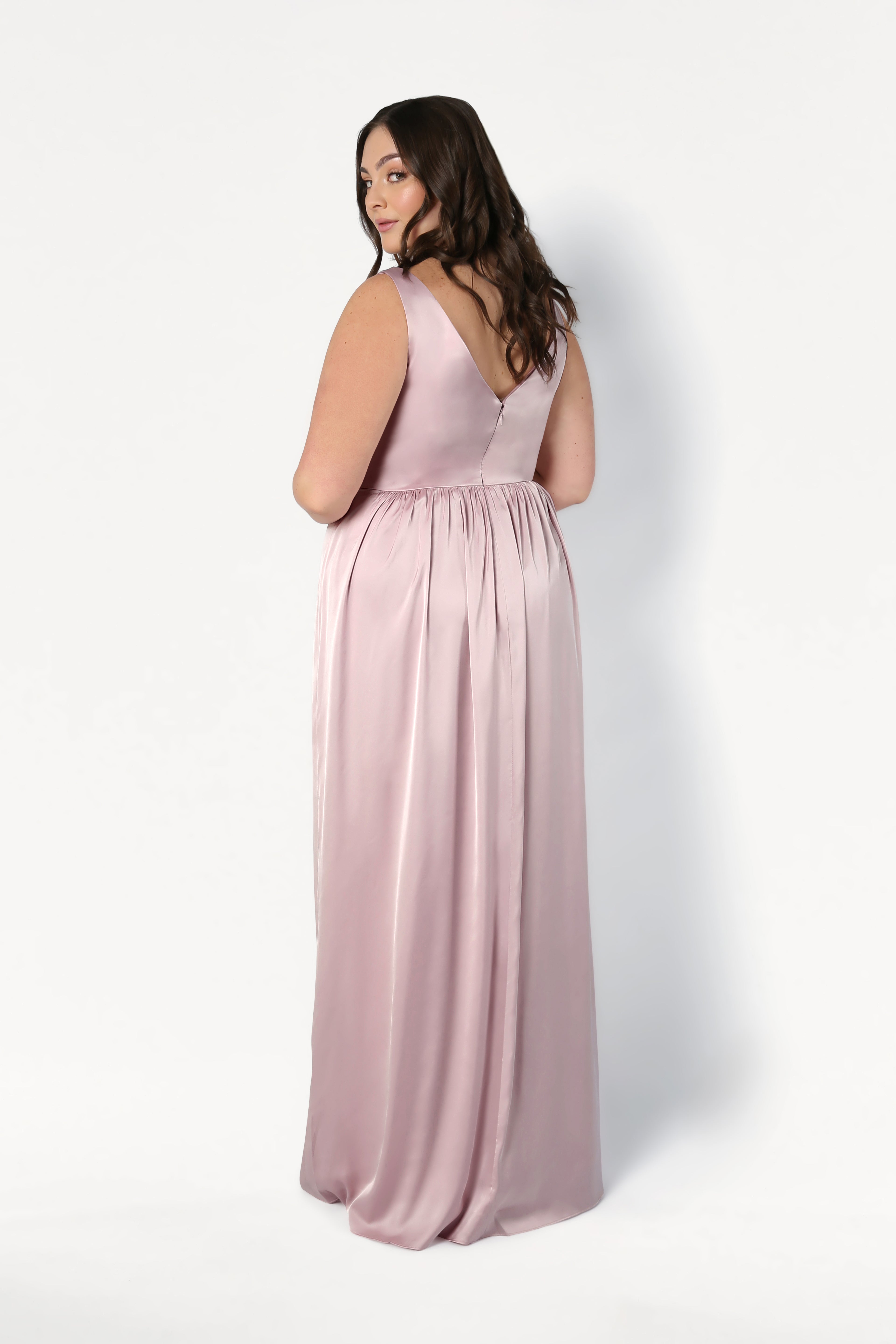 A woman with long brown hair models the TH&TH Carmen Bridesmaid Dress in Smoked Blush Satin—a sleeveless, floor-length gown—standing against a white background and looking over her shoulder; a chic choice for modern bridal parties.