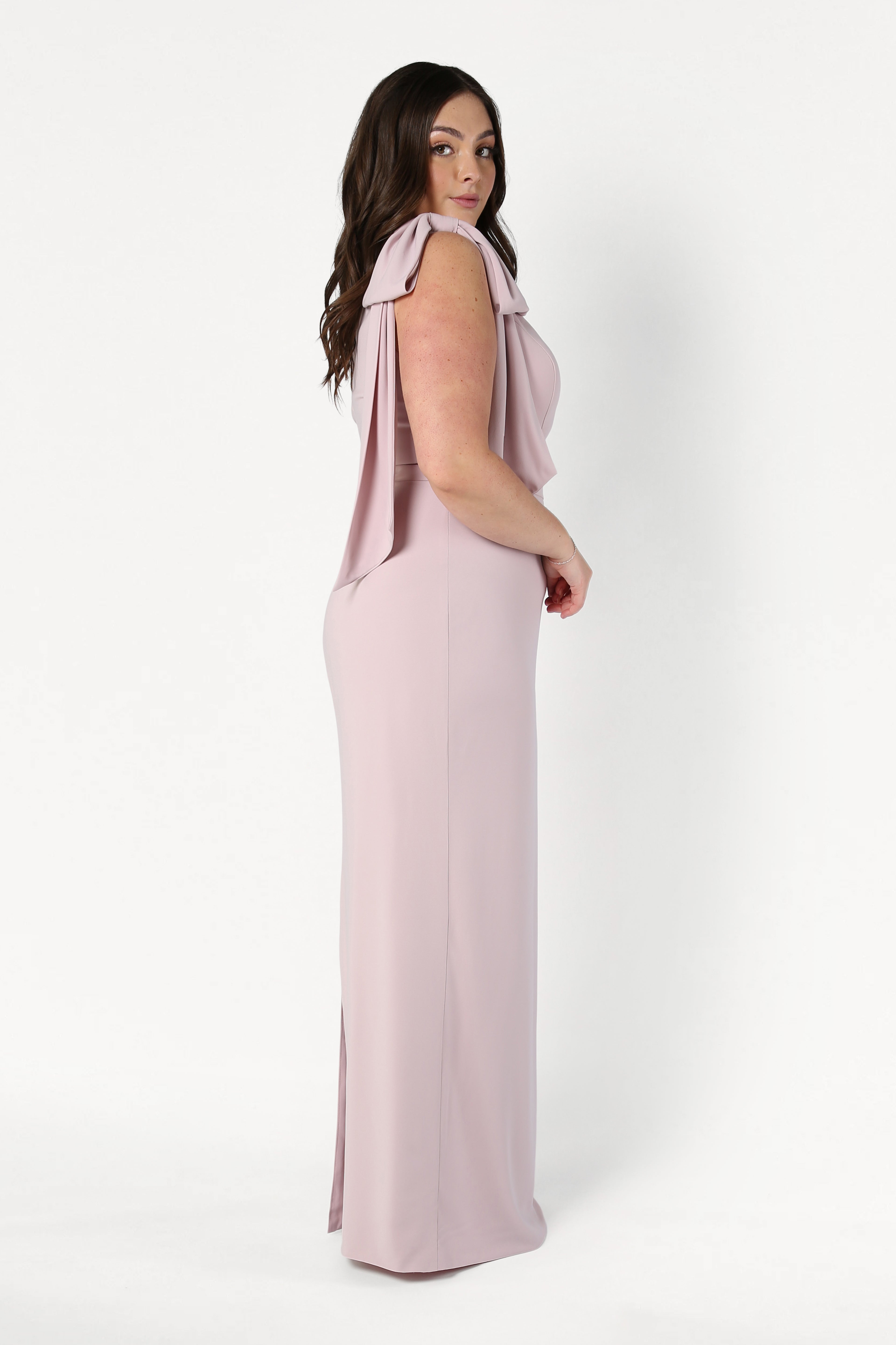 A woman with long dark hair stands in profile against a white background, wearing the TH&TH Valentina Bridesmaid Dress in Smoked Blush—a floor-length, sleeveless gown featuring draped shoulder details.