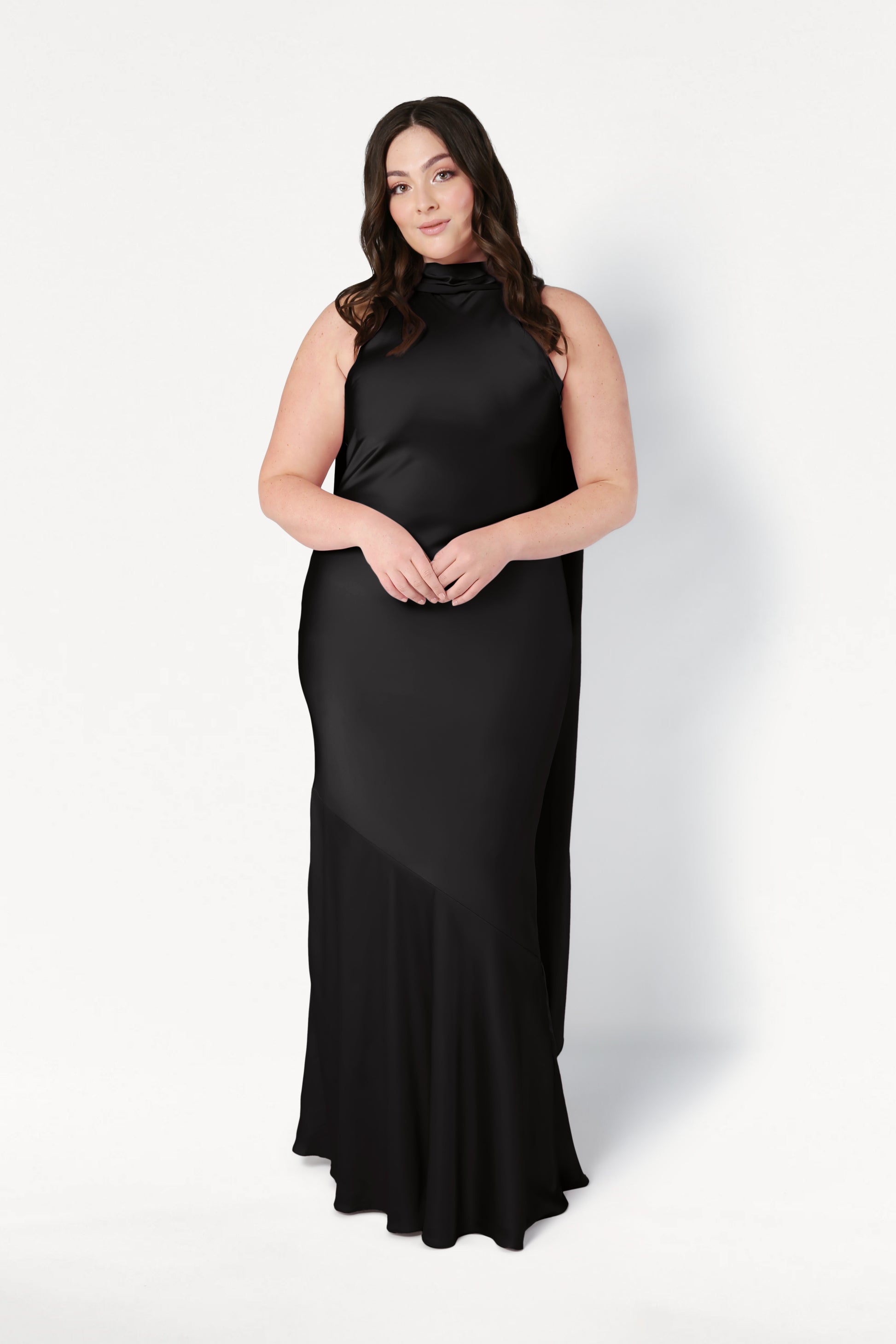 A woman with long brown hair wears the Mira Bridesmaid Dress in Black Satin by TH&TH. She stands against a plain white background, facing forward with her hands gently clasped in front of her.