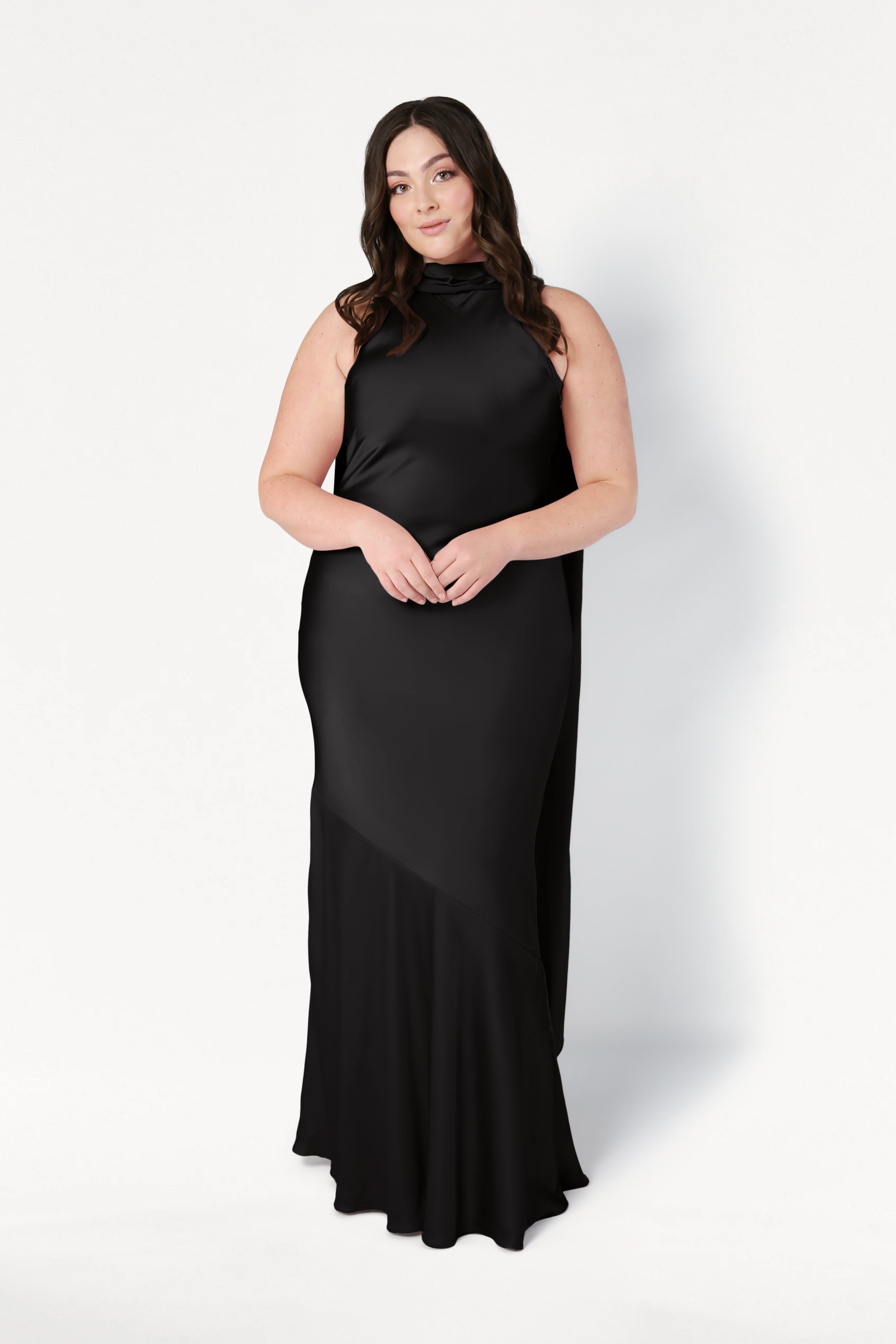 A woman with long brown hair wears the Mira Bridesmaid Dress in Black Satin by TH&TH. She stands against a plain white background, facing forward with her hands gently clasped in front of her.