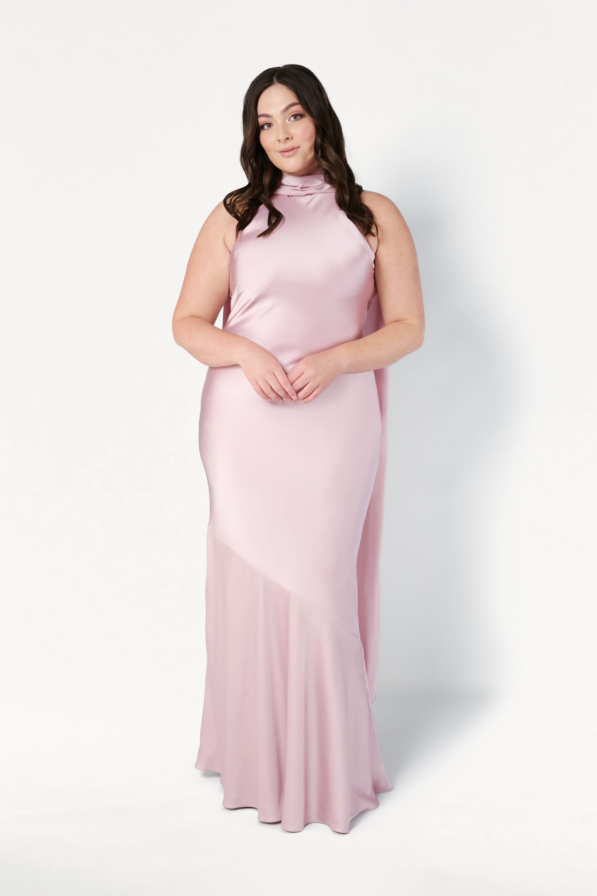 A woman with long dark hair wears the TH&TH Mira Bridesmaid Dress in blush pink satin, featuring a sleeveless design and high neckline, standing against a plain white background.