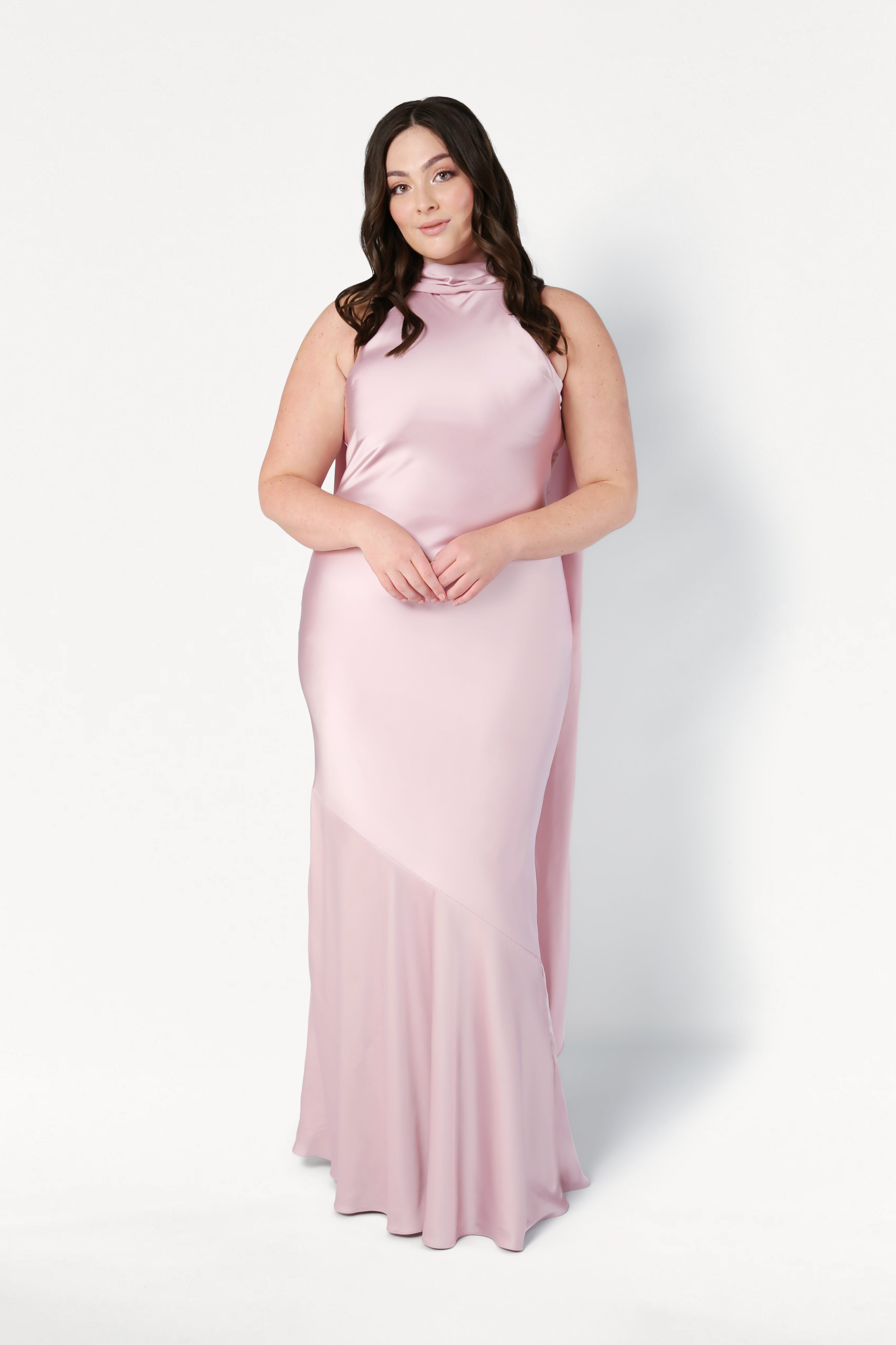 A woman with long dark hair wears the TH&TH Mira Bridesmaid Dress in blush pink satin, featuring a sleeveless design and high neckline, standing against a plain white background.