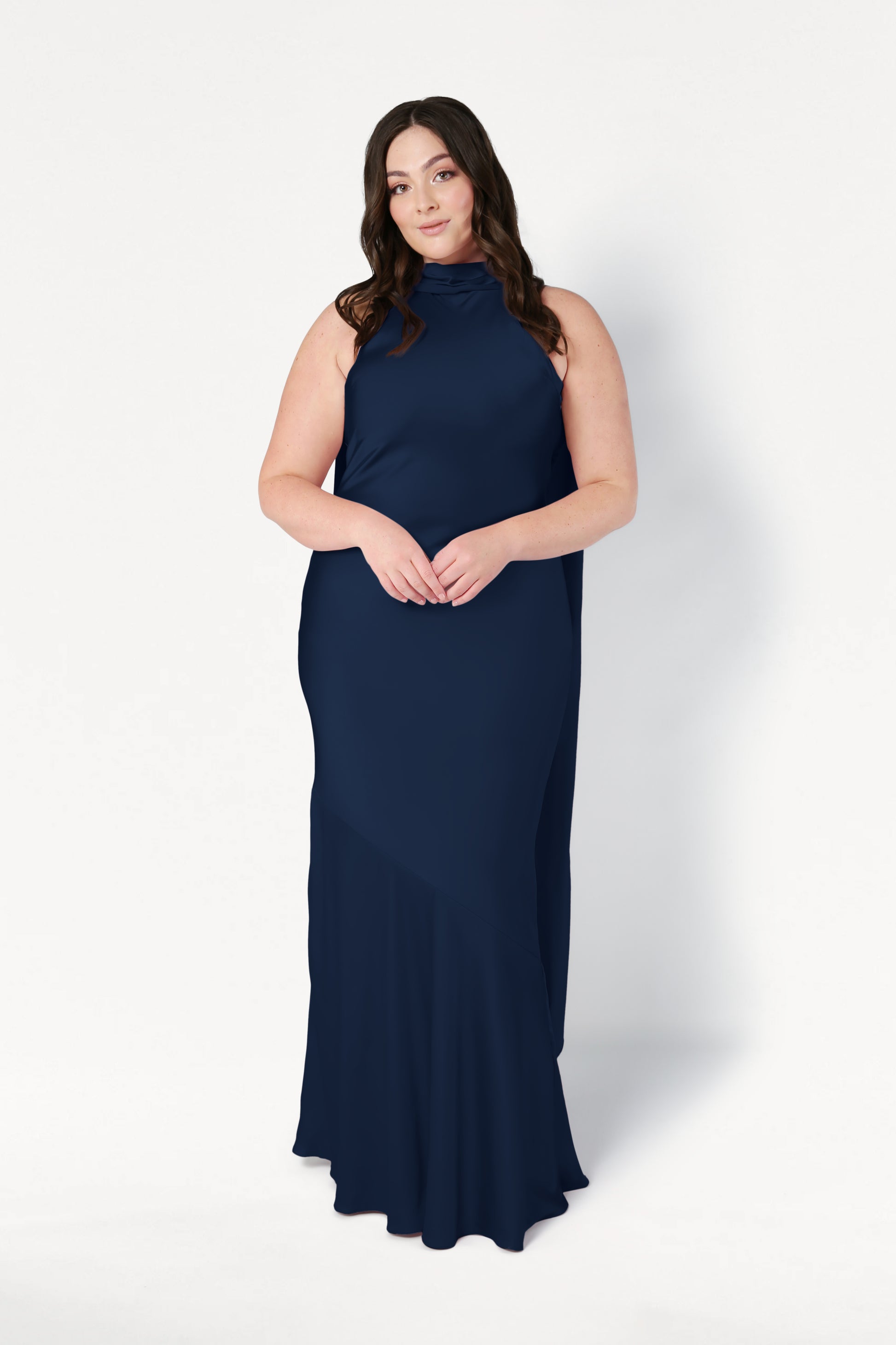 A woman with long brown hair stands against a white background, hands clasped, wearing the TH&TH Mira Bridesmaid Dress in navy satin—a sleeveless, floor-length gown.