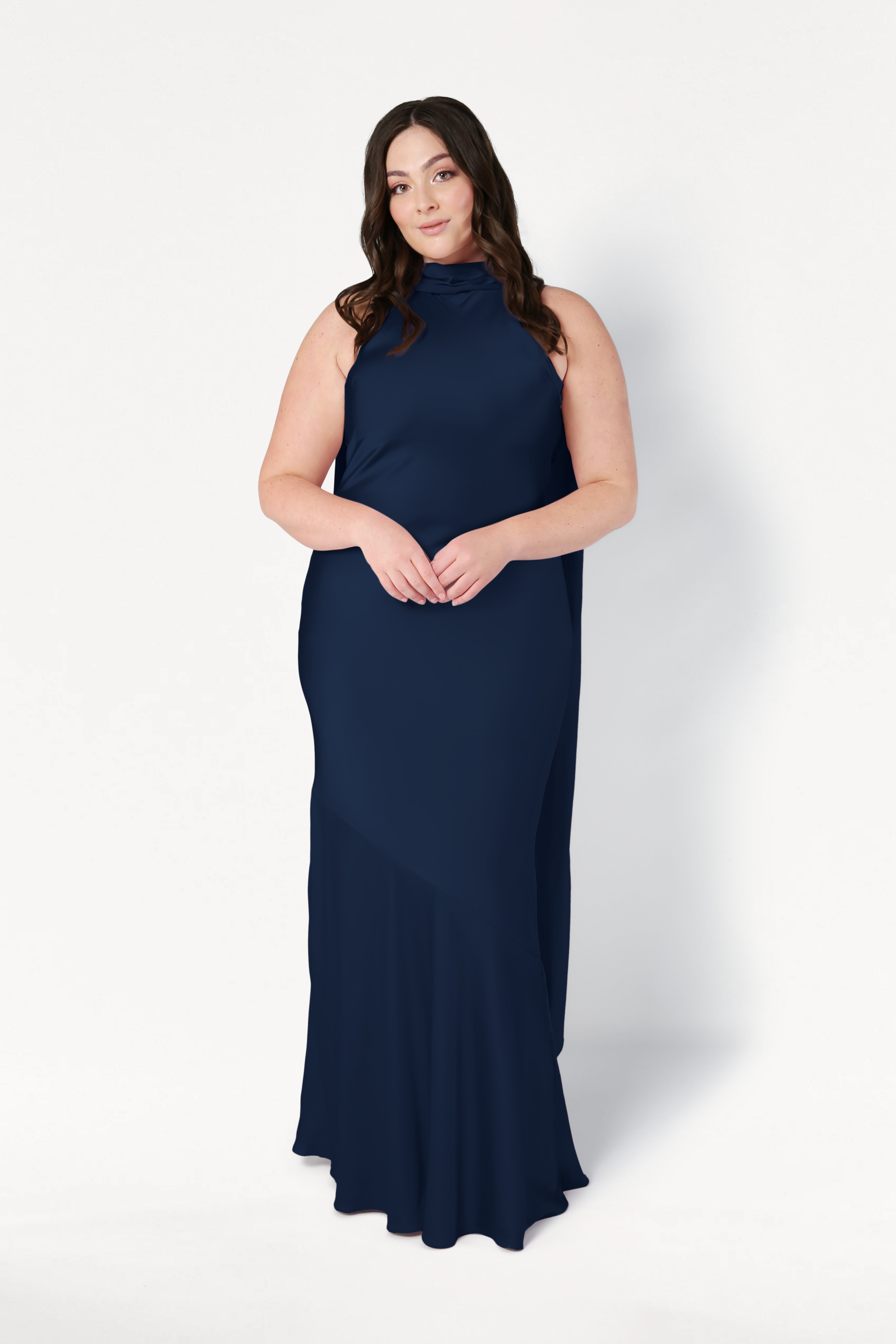 A woman with long brown hair stands against a white background, hands clasped, wearing the TH&TH Mira Bridesmaid Dress in navy satin—a sleeveless, floor-length gown.