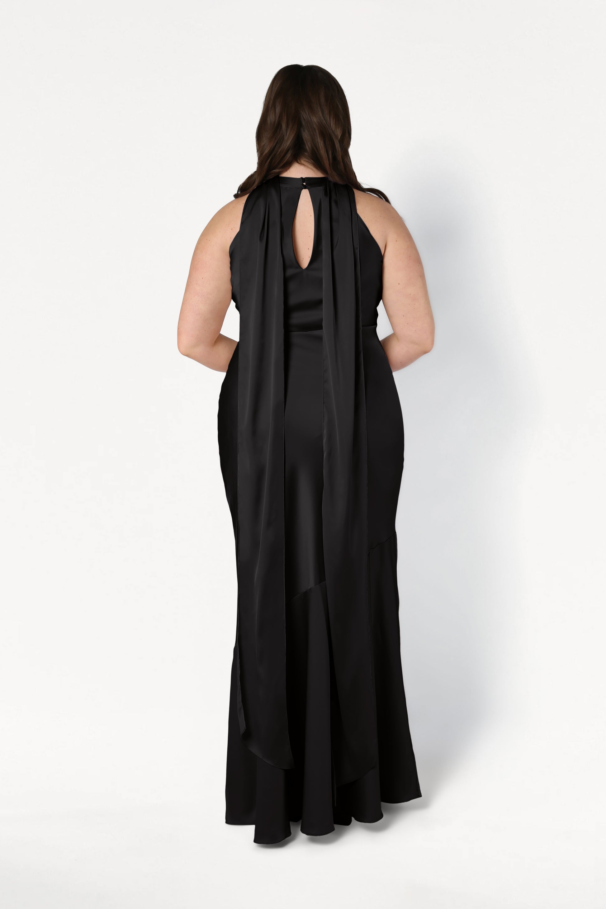 A woman wears the TH&TH Mira Bridesmaid Dress in black satin, showing its long, sleeveless design with a keyhole back and flowing shoulder panels as she poses facing away against a plain white background.