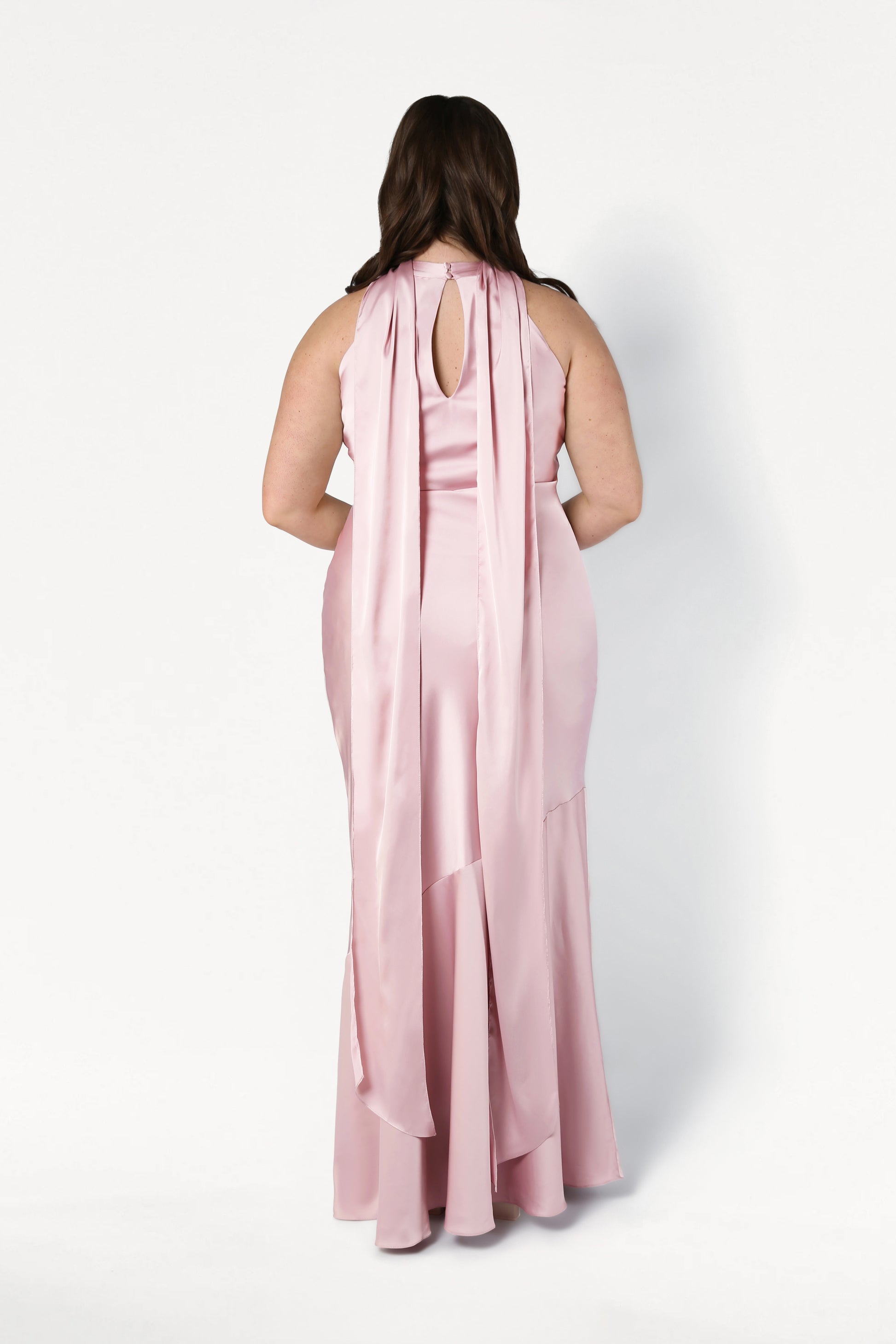 A woman with long brown hair models the TH&TH Mira Bridesmaid Dress in Blush Pink Satin, showcasing its sleeveless, floor-length bias-cut silhouette, keyhole back detail, and flowing panels against a plain white backdrop.