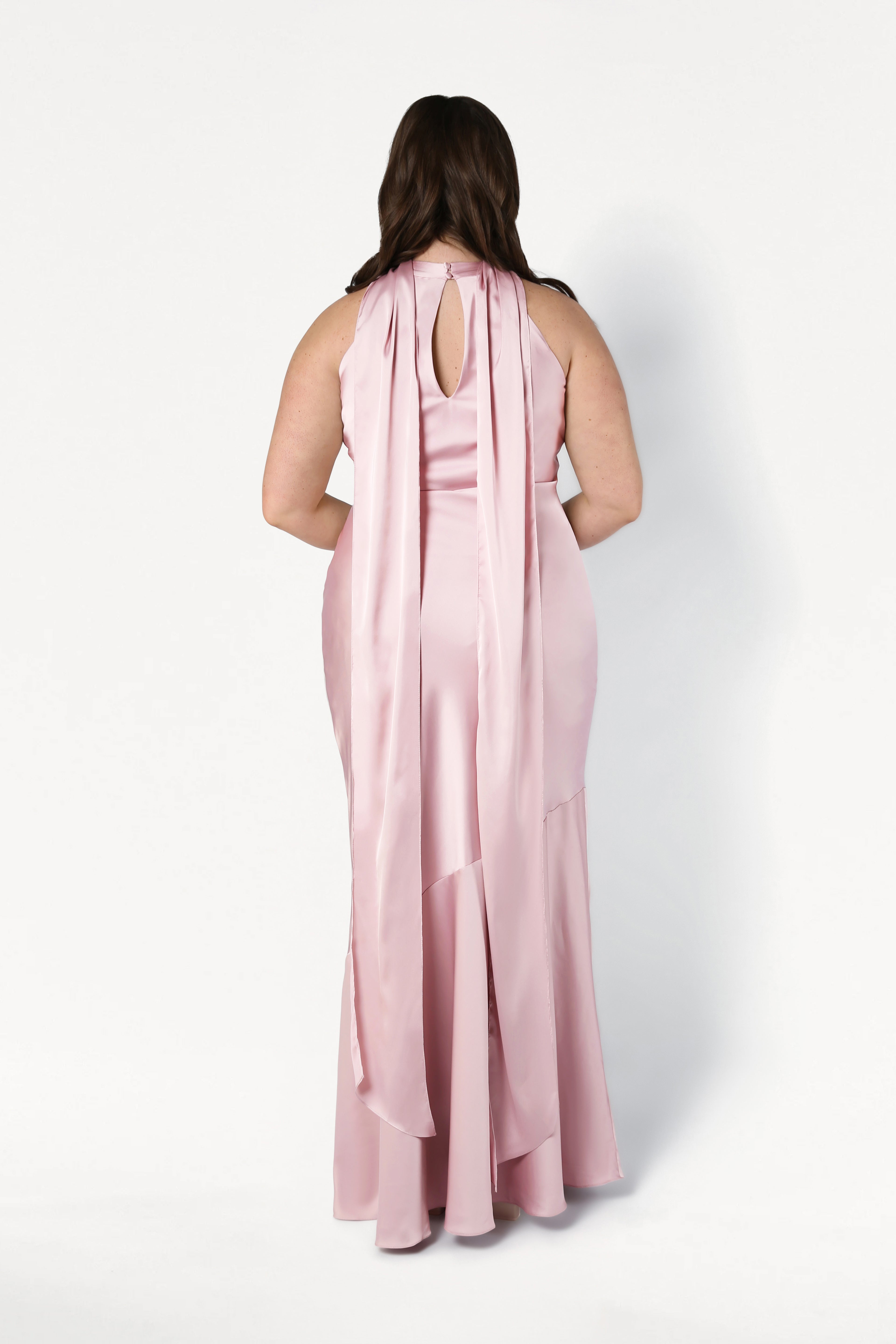 A woman with long brown hair models the TH&TH Mira Bridesmaid Dress in Blush Pink Satin, showcasing its sleeveless, floor-length bias-cut silhouette, keyhole back detail, and flowing panels against a plain white backdrop.
