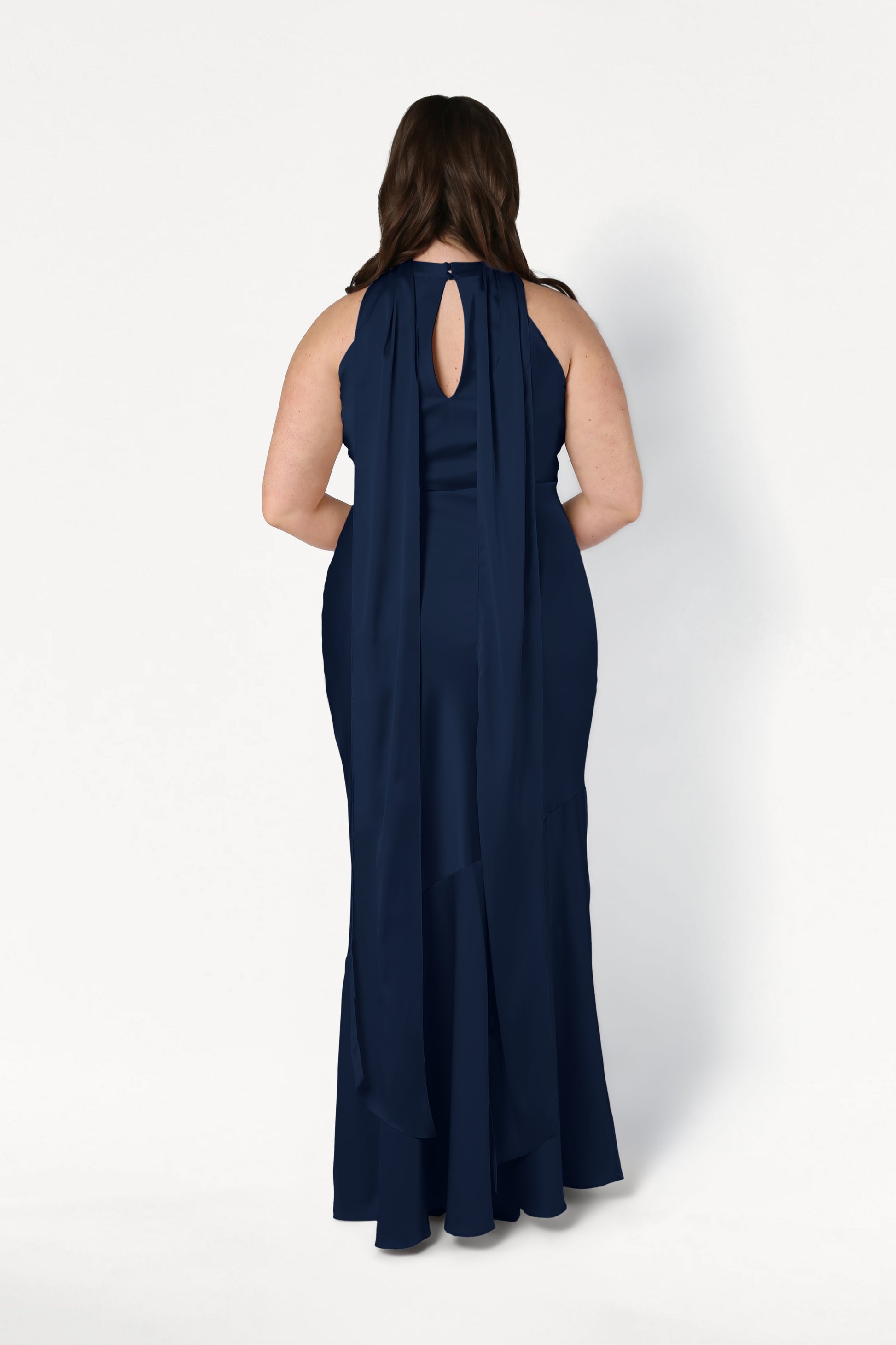 A woman stands facing away against a plain white background, wearing the TH&TH Mira Bridesmaid Dress in Navy Satin—a floor-length halterneck dress with a keyhole back and long, flowing scarf-like details.