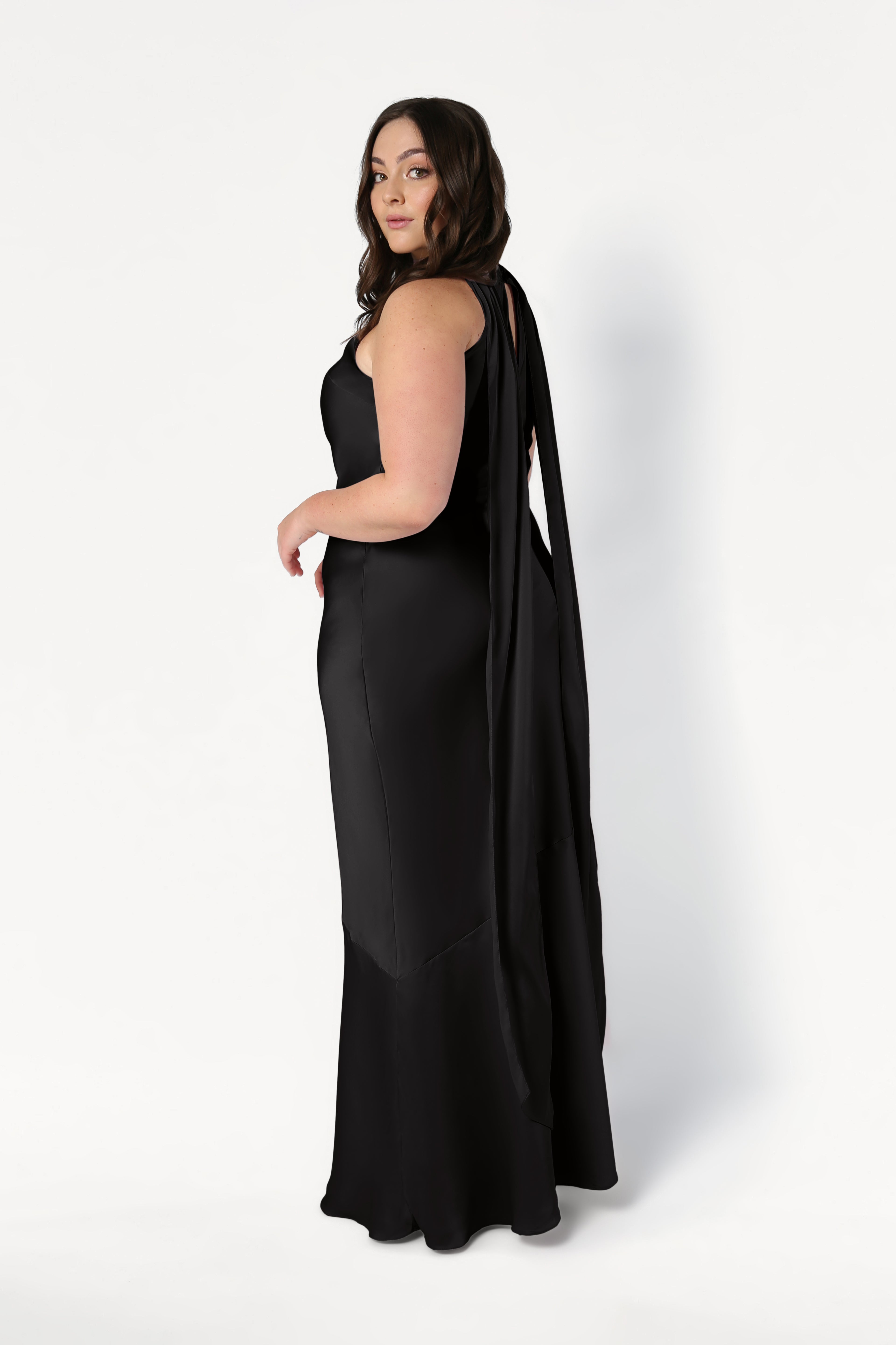 A woman with long brown hair models the TH&TH Mira Bridesmaid Dress in Black Satin, featuring a draped sash on the back, as she stands sideways and looks over her shoulder against a plain white background.