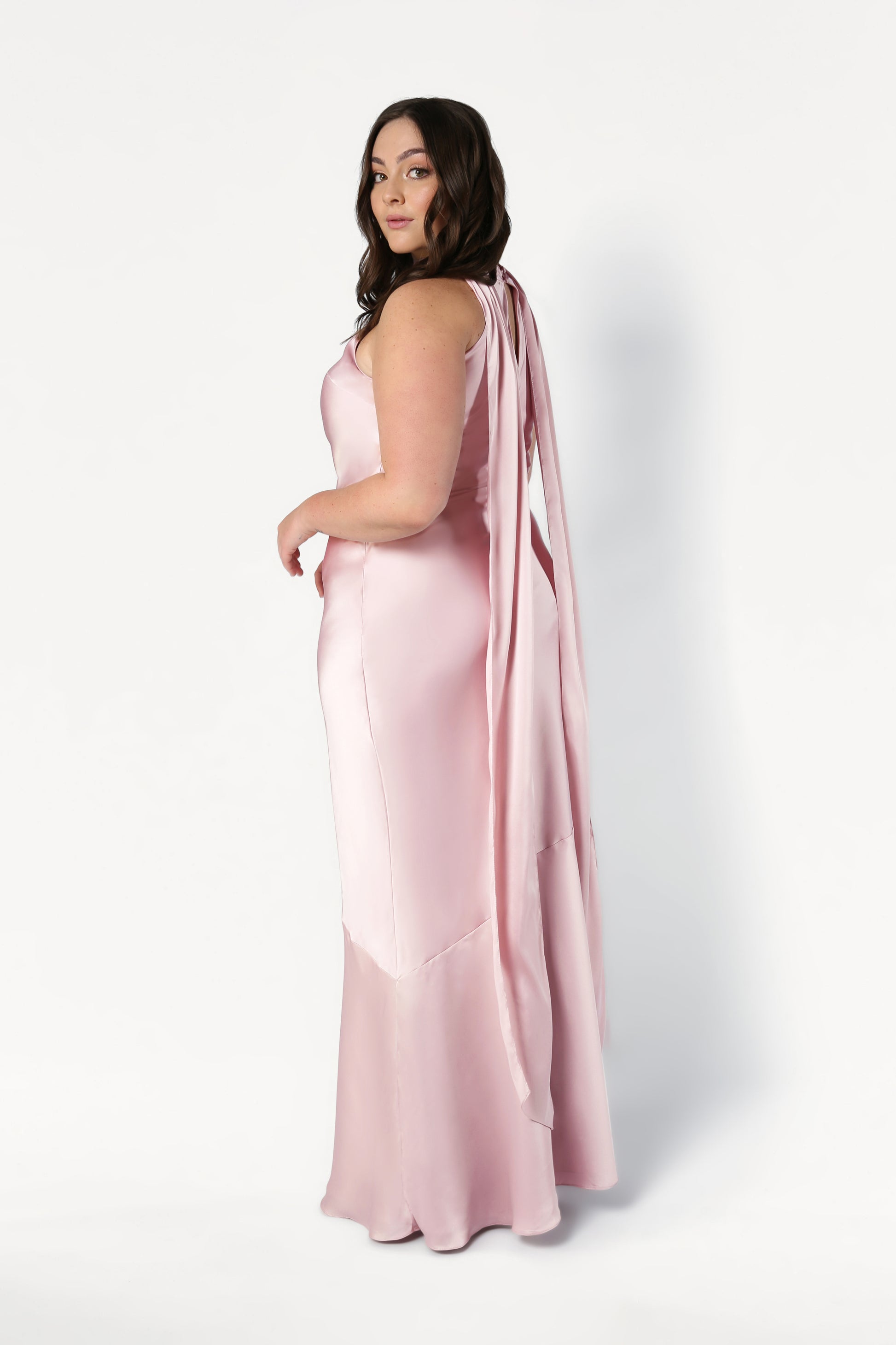 A woman with long brown hair poses sideways against a white background wearing the TH&TH Mira Bridesmaid Dress in blush pink satin, featuring a floor-length silhouette and long ribbon details.