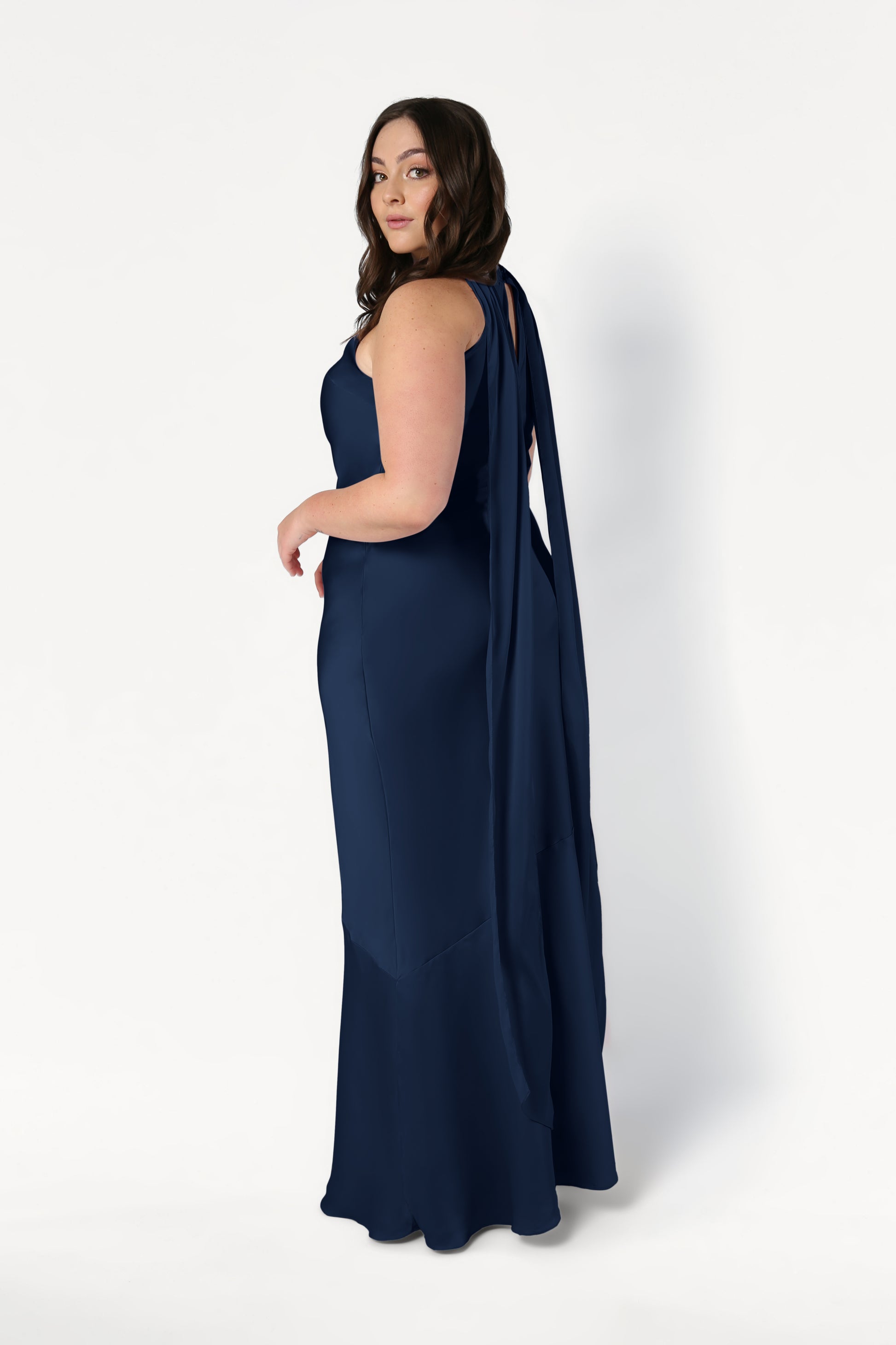 A woman with wavy brown hair models the TH&TH Mira Bridesmaid Dress in Navy Satin, featuring a sleeveless, floor-length design and draped back detail, posed slightly to the side against a white background.