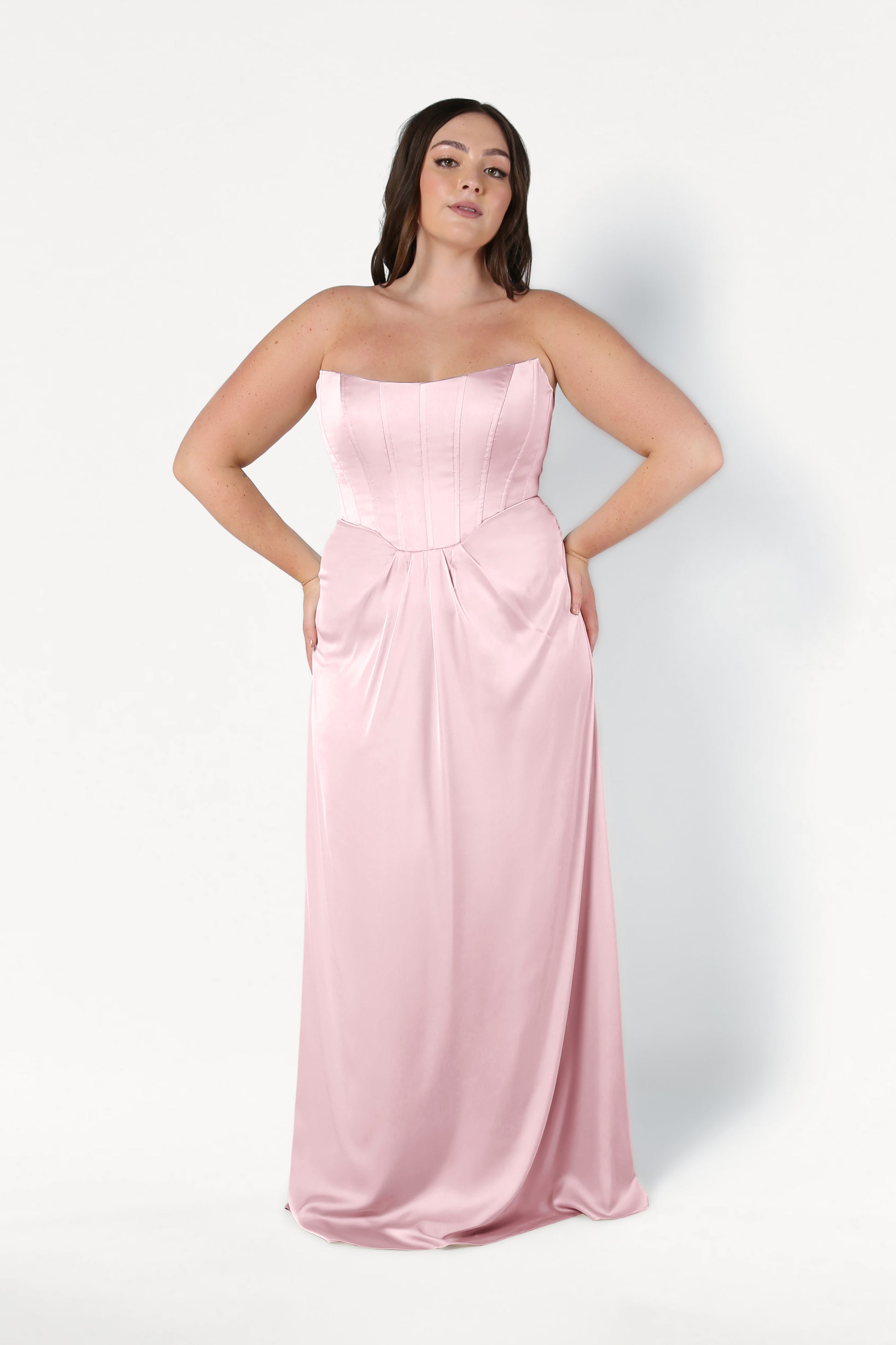 A woman with long dark hair poses against a white background wearing the TH&TH Selena Bridesmaid Dress in blush pink satin, featuring a strapless fitted corset bodice and flowing skirt, with her hands on her hips.