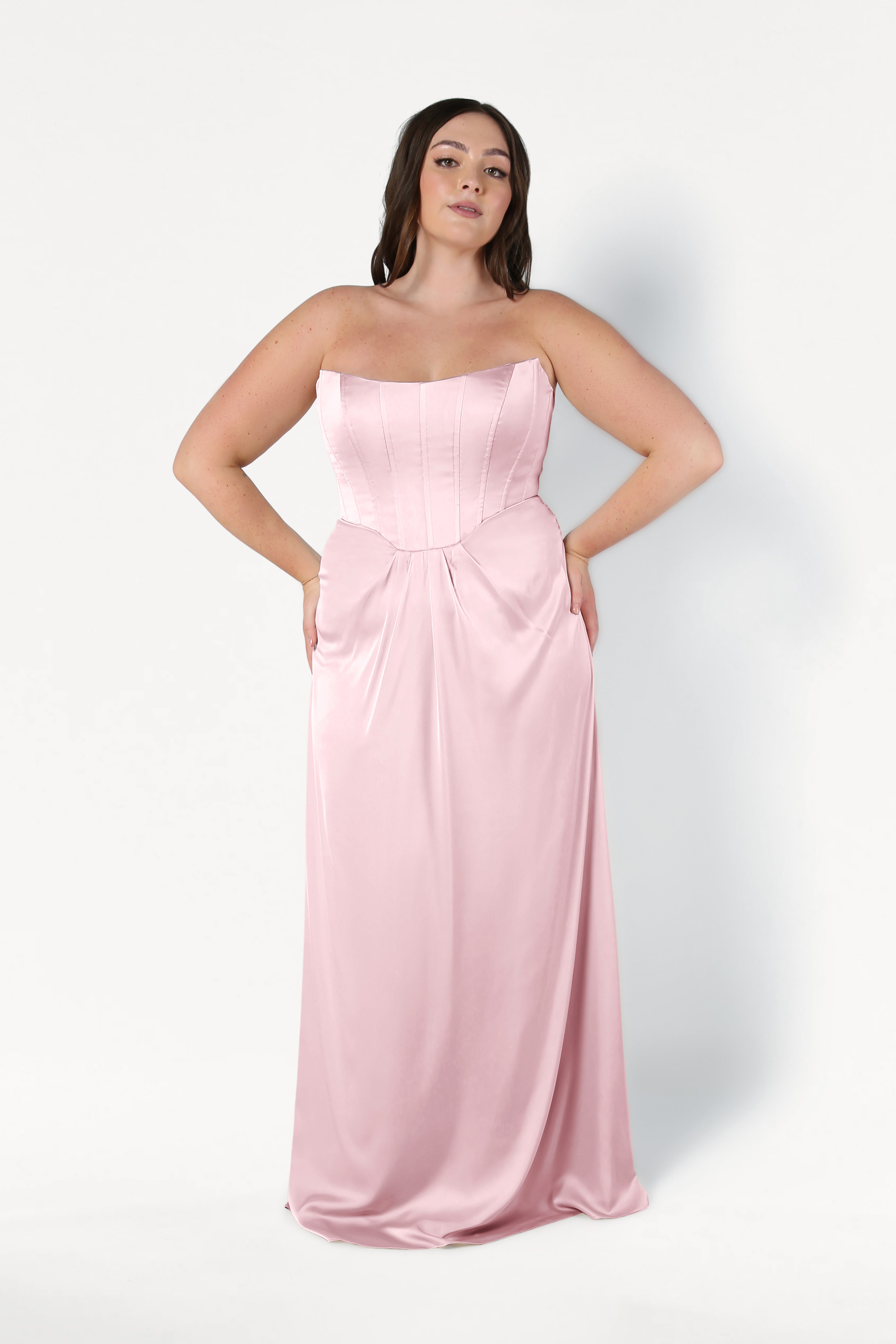 A woman with long dark hair poses against a white background wearing the TH&TH Selena Bridesmaid Dress in blush pink satin, featuring a strapless fitted corset bodice and flowing skirt, with her hands on her hips.