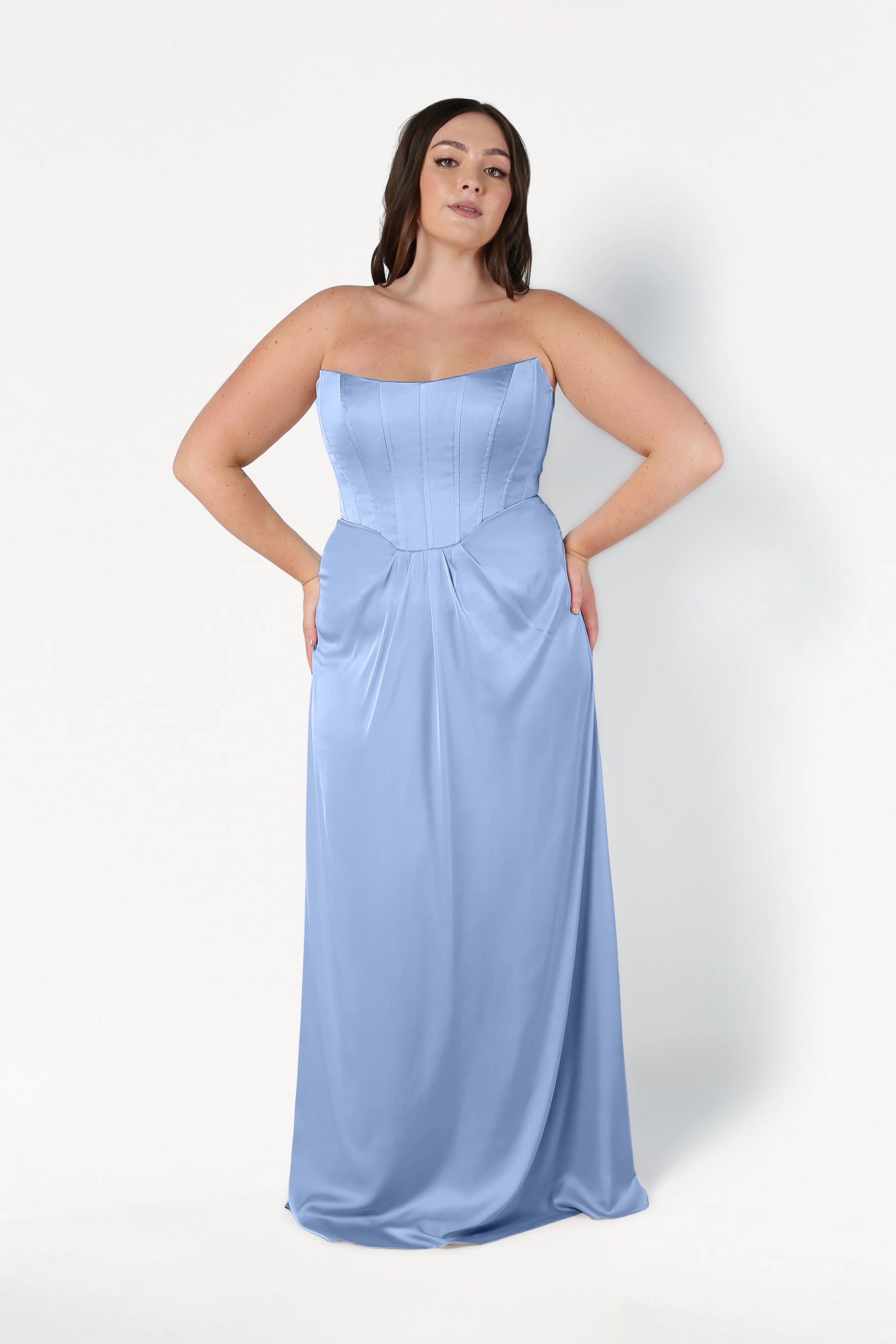 A woman poses confidently against a white background wearing the TH&TH Selena Bridesmaid Dress in Dusty Blue Satin, featuring a size-inclusive fit, fitted corset bodice, and flowing skirt. Her hands are on her hips as she faces the camera.