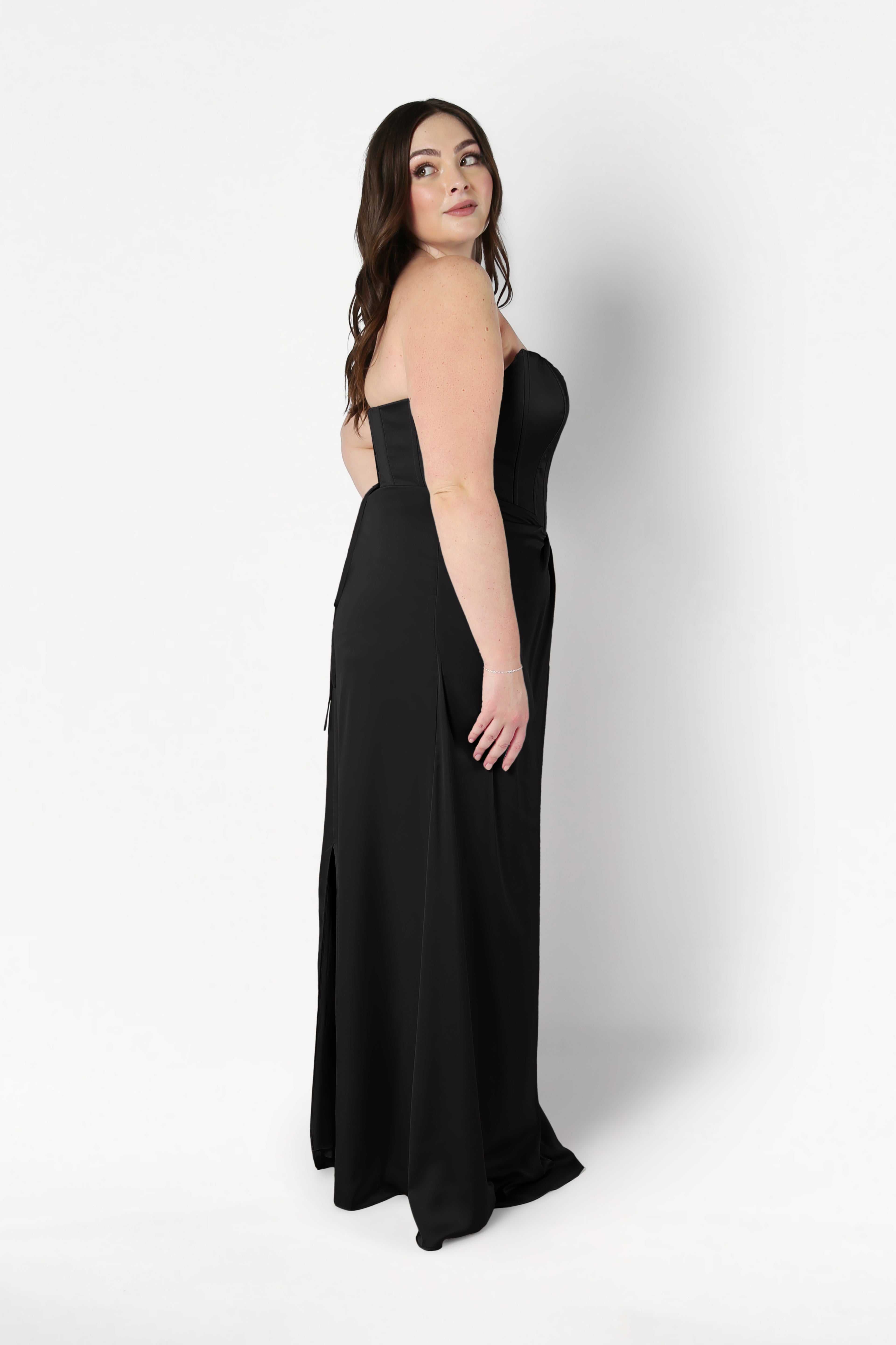 Woman with long brown hair stands sideways against a plain white background, looking over her left shoulder, wearing the TH&TH Selena Bridesmaid Dress in Black Satin with a strapless corset bodice and floor-length silhouette.